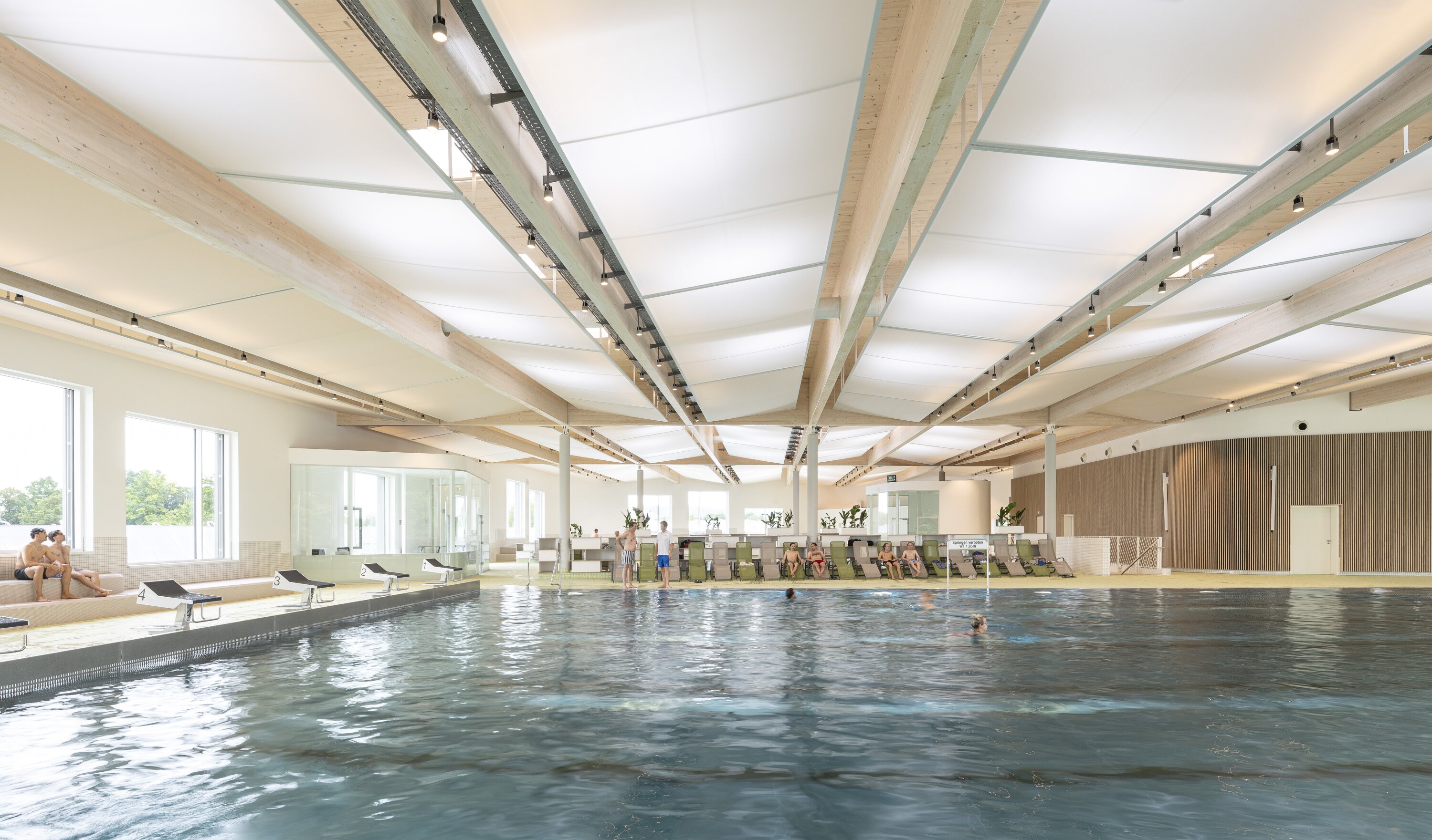 Project by Behnisch Architekturbuero, Sportpark Ost Regensburg. Indoor swimming pool with a seating area for visitors, large windows, and a wooden beamed ceiling. People are swimming in the large pool. Starting blocks can be seen on the left side of the pool.