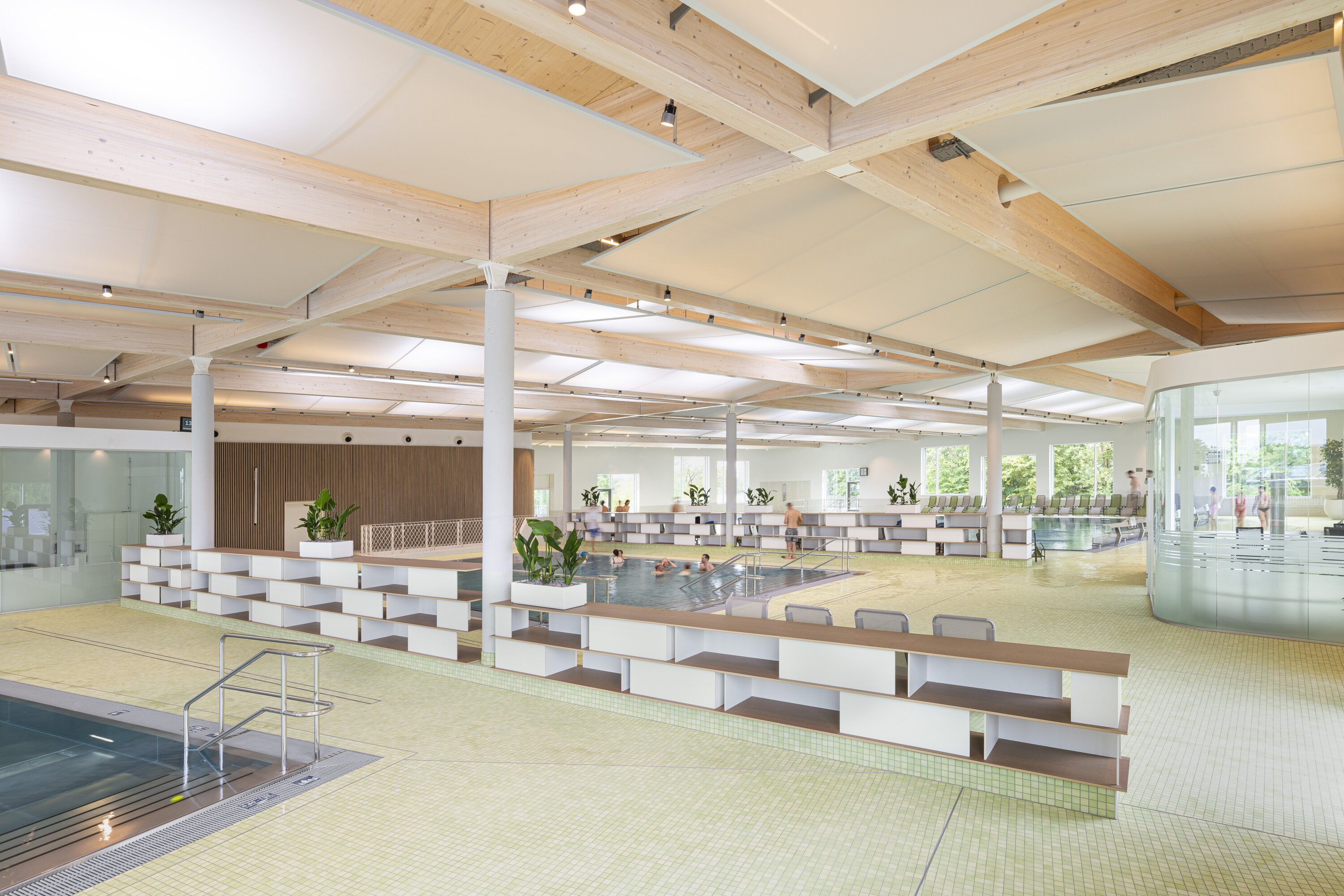 Project by Behnisch Architekturbuero, Sportpark Ost Regensburg. Indoor swimming pool hall with bathers, seating, and wood ceiling structure. The lifeguards' room is visible on the right.
