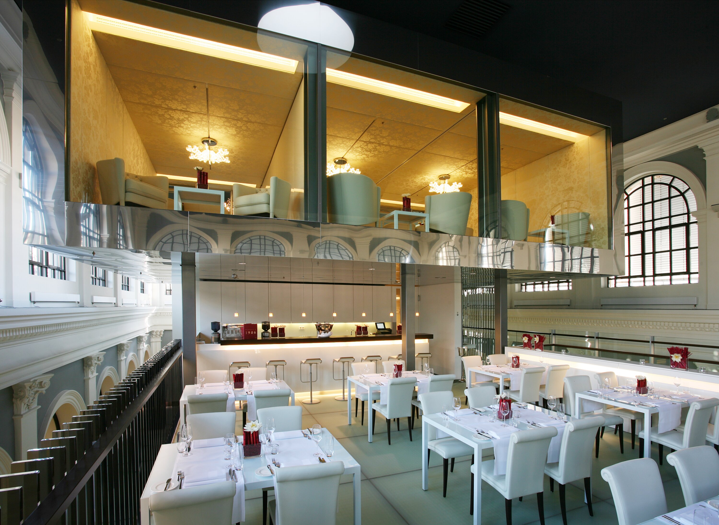 Project by Behnisch Architekturbuero, Haus im Haus. Project by Behnisch Architekturbuero, Haus im Haus. Modern restaurant interior with white tables and chairs, bar counter, and a glass-walled lounge above within a historic hall.