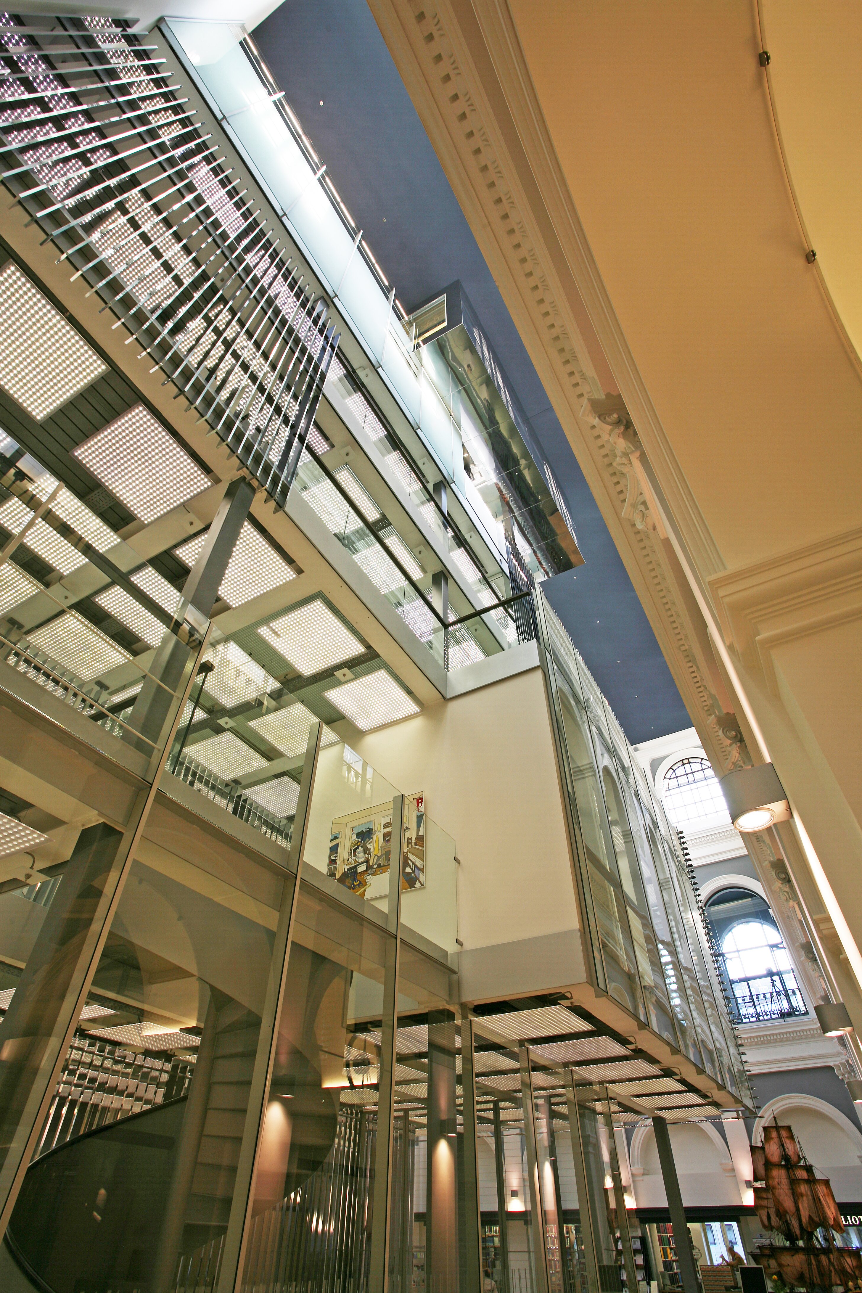 Project by Behnisch Architekturbuero, Haus im Haus. Upward view of modern glass mezzanine inserted into historic hall, with gridded ceiling panels, columns, and arched windows visible.
