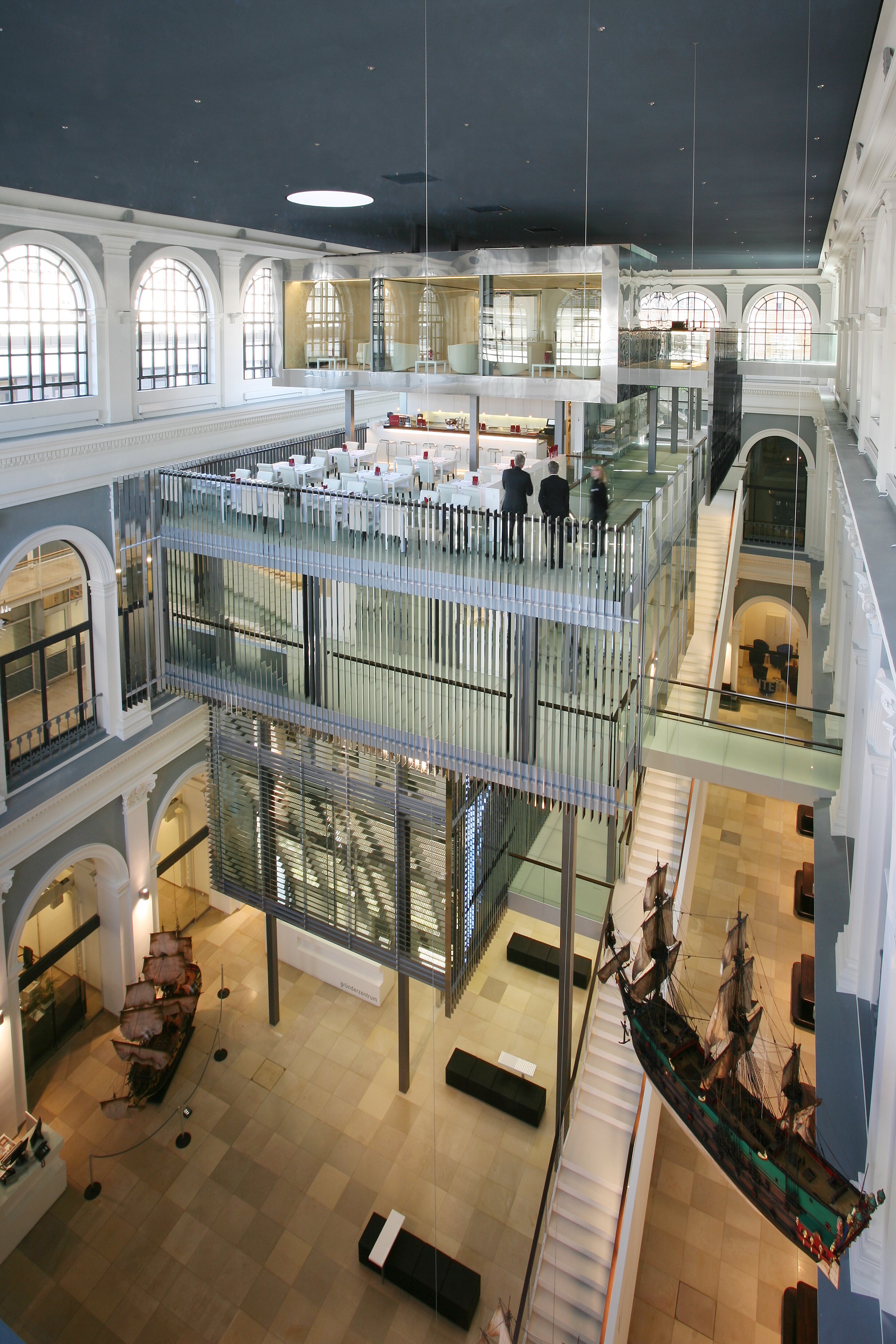 Project by Behnisch Architekturbuero, Haus im Haus. Multi-level atrium with glass cubes, dining area above, staircases, and historic hall with ship models displayed below.