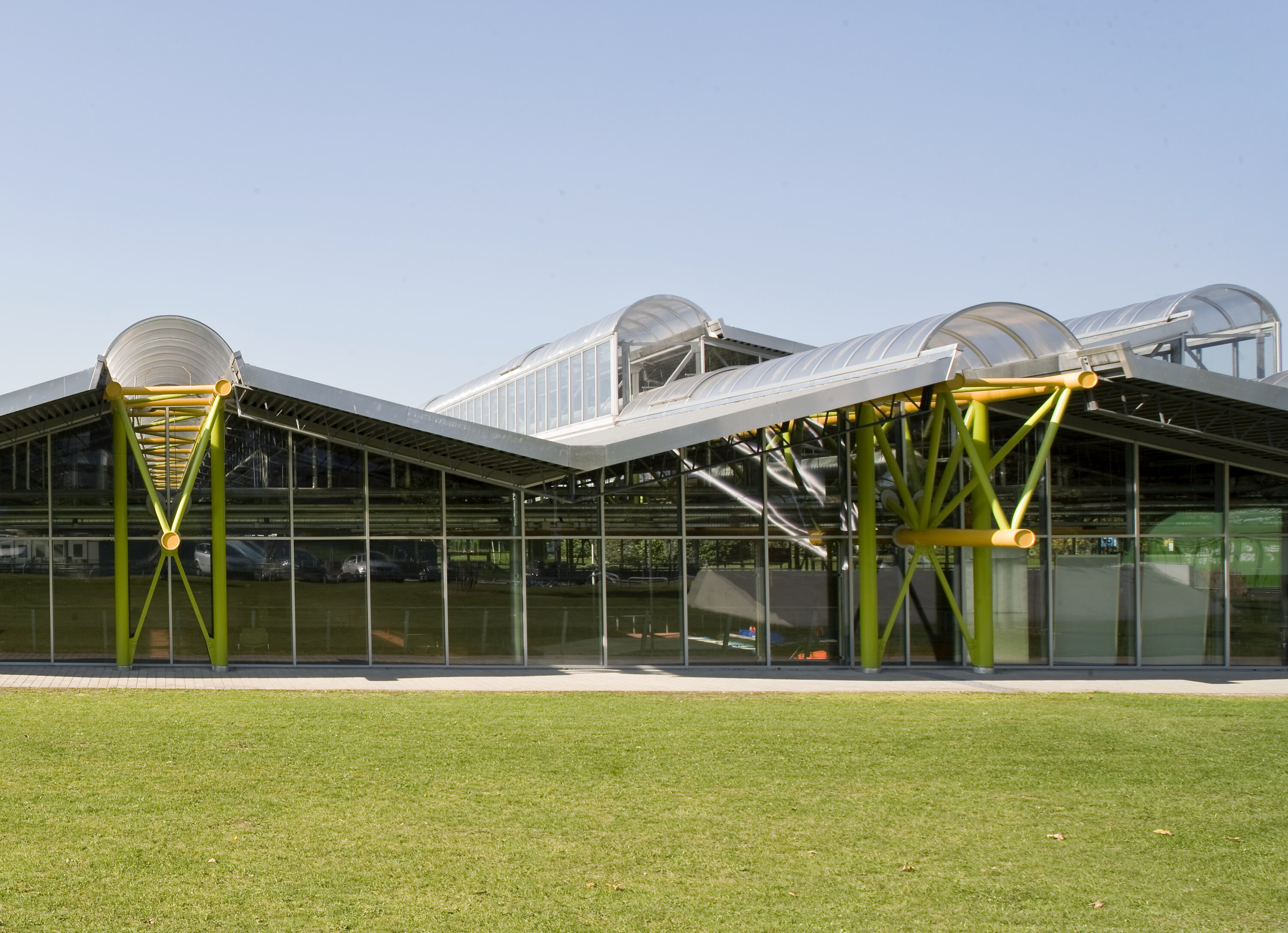 Project by Behnisch Architekturbuero, Werner-von-Linde-Halle. Modern glass building with curved roof sections and green steel supports, viewed from a grassy foreground under a clear sky.