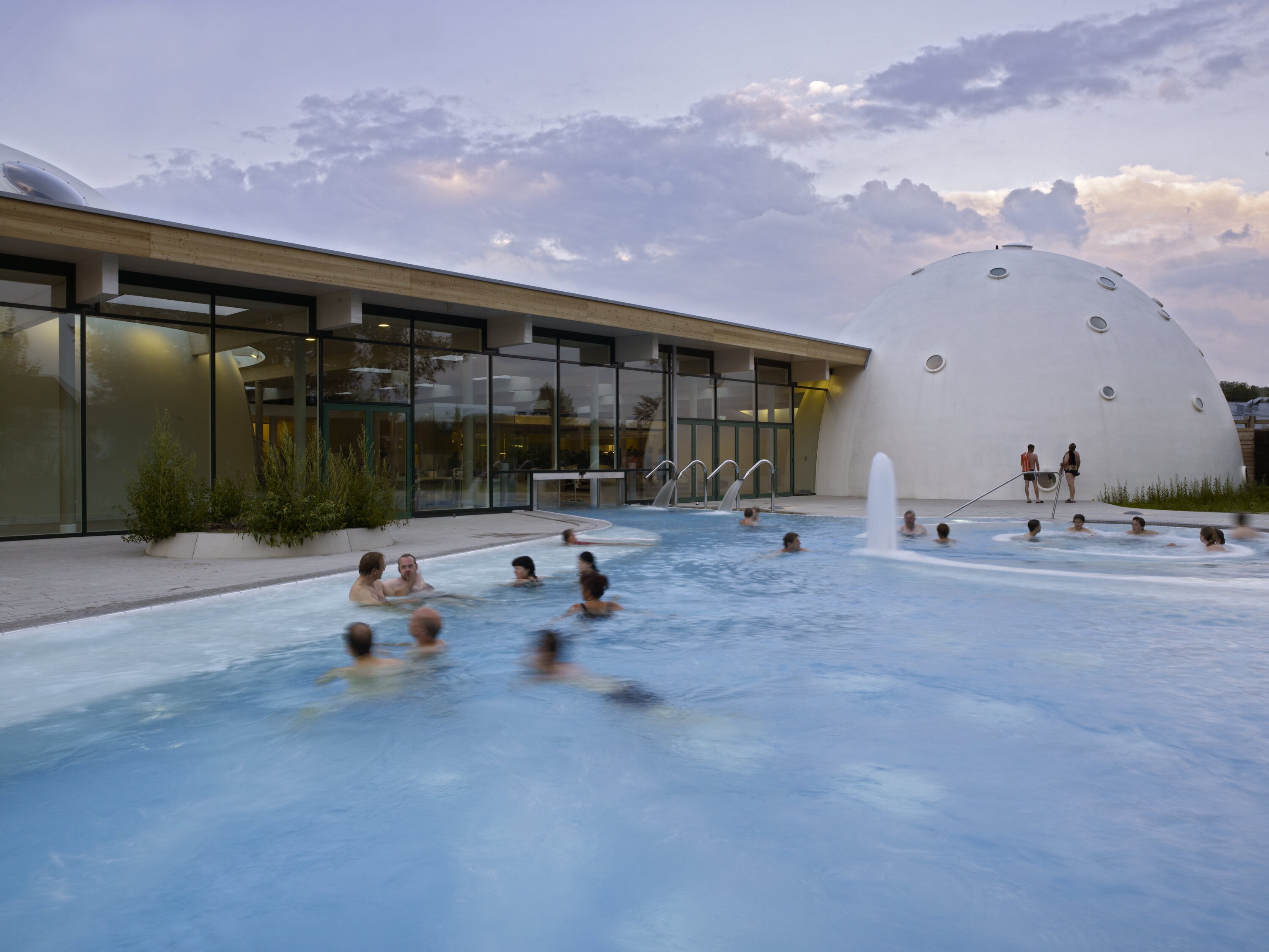 Project by Behnisch Architekturbuero, Thermal Spa Bad Aibling. Outdoor pool with people bathing beside the modern glass building and a white domed structure, with water jets and evening sky in the background.