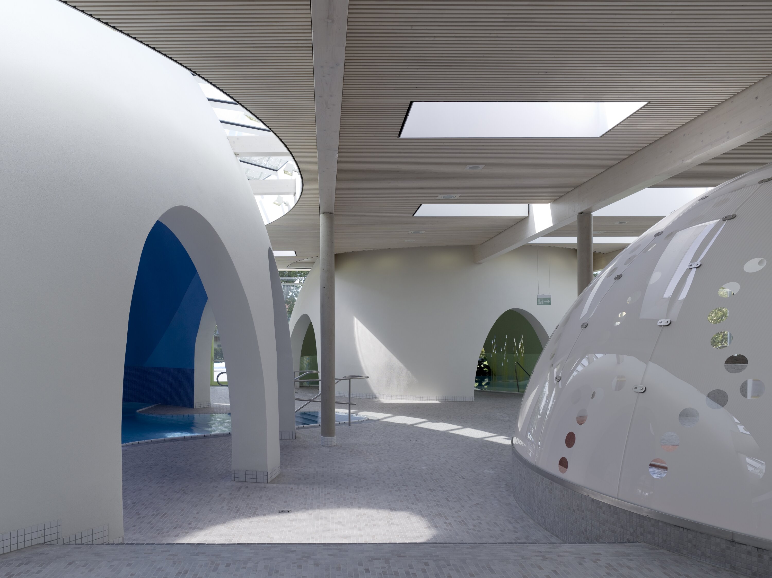 Project by Behnisch Architekturbuero, Thermal Spa Bad Aibling. Indoor pool area with white curved structures, arched openings, skylights, and tiled floors, with small pools visible beneath the arches.