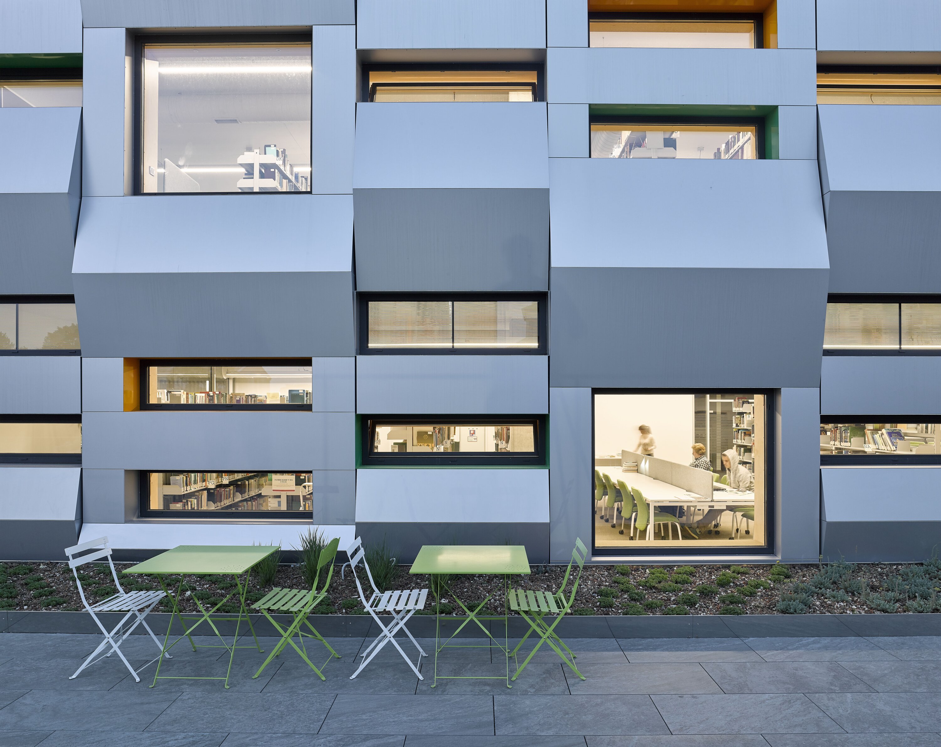 Project by Behnisch Architekturbuero, The Metropolitan Sheptytsky Center, Ukrainian Catholic University Lviv. A modern building facade with windows of varying sizes, offering a view into the library's interior. Tables and chairs are set up on a terrace in front.