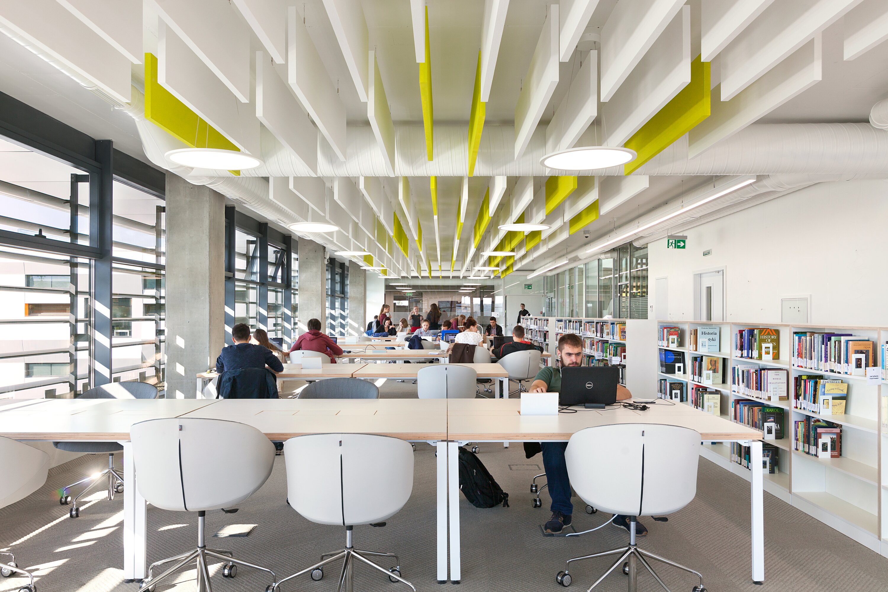 Project by Behnisch Architekturbuero, The Metropolitan Sheptytsky Center, Ukrainian Catholic University Lviv. Library reading room with long tables, bookshelves, and people studying on laptops under white and yellow ceiling panels.