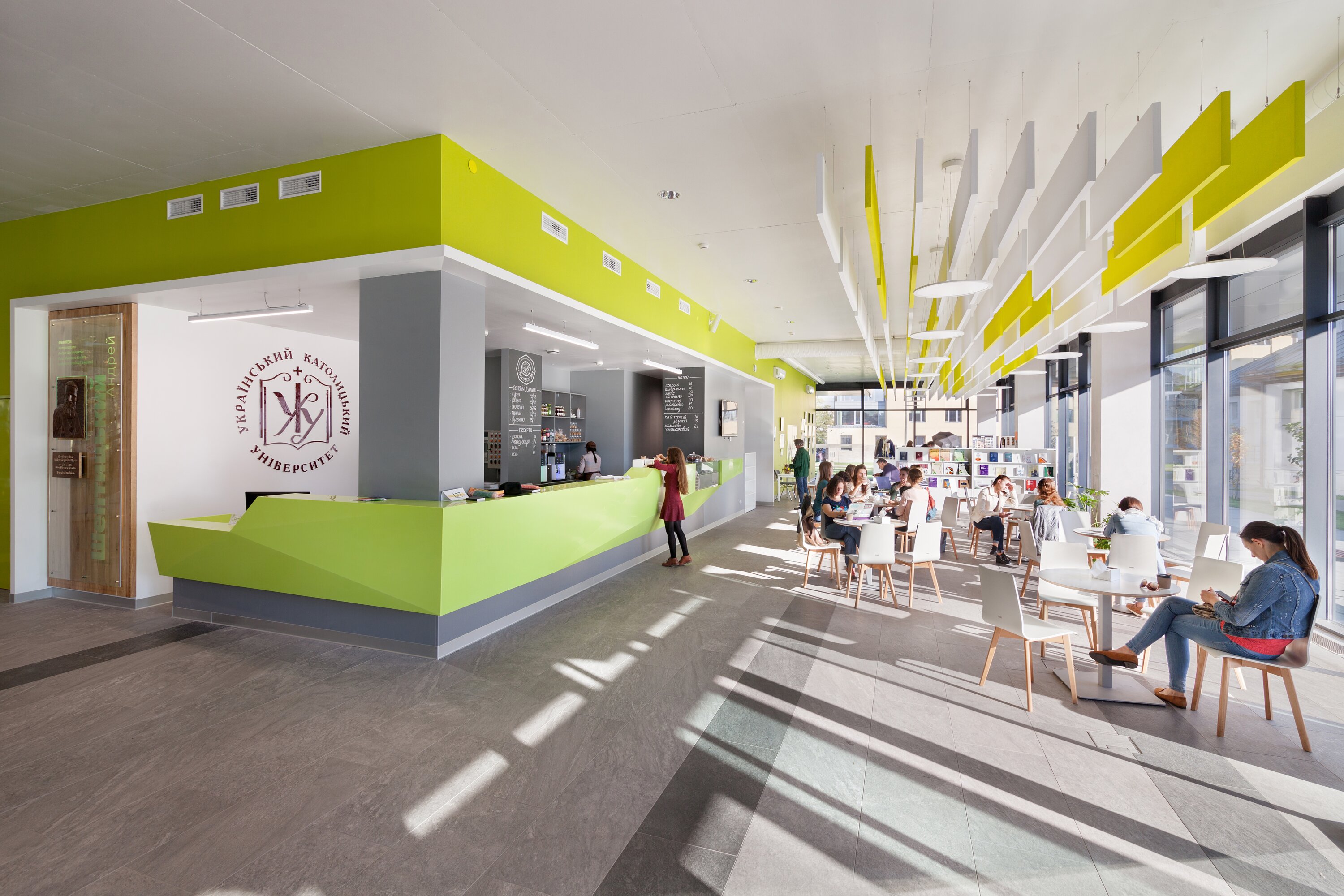 Project by Behnisch Architekturbuero, The Metropolitan Sheptytsky Center, Ukrainian Catholic University Lviv. Bright caf&eacute; interior with a green counter, students at tables, and a glass facade that lets in daylight. White and yellow acoustic panels hang from the ceiling.