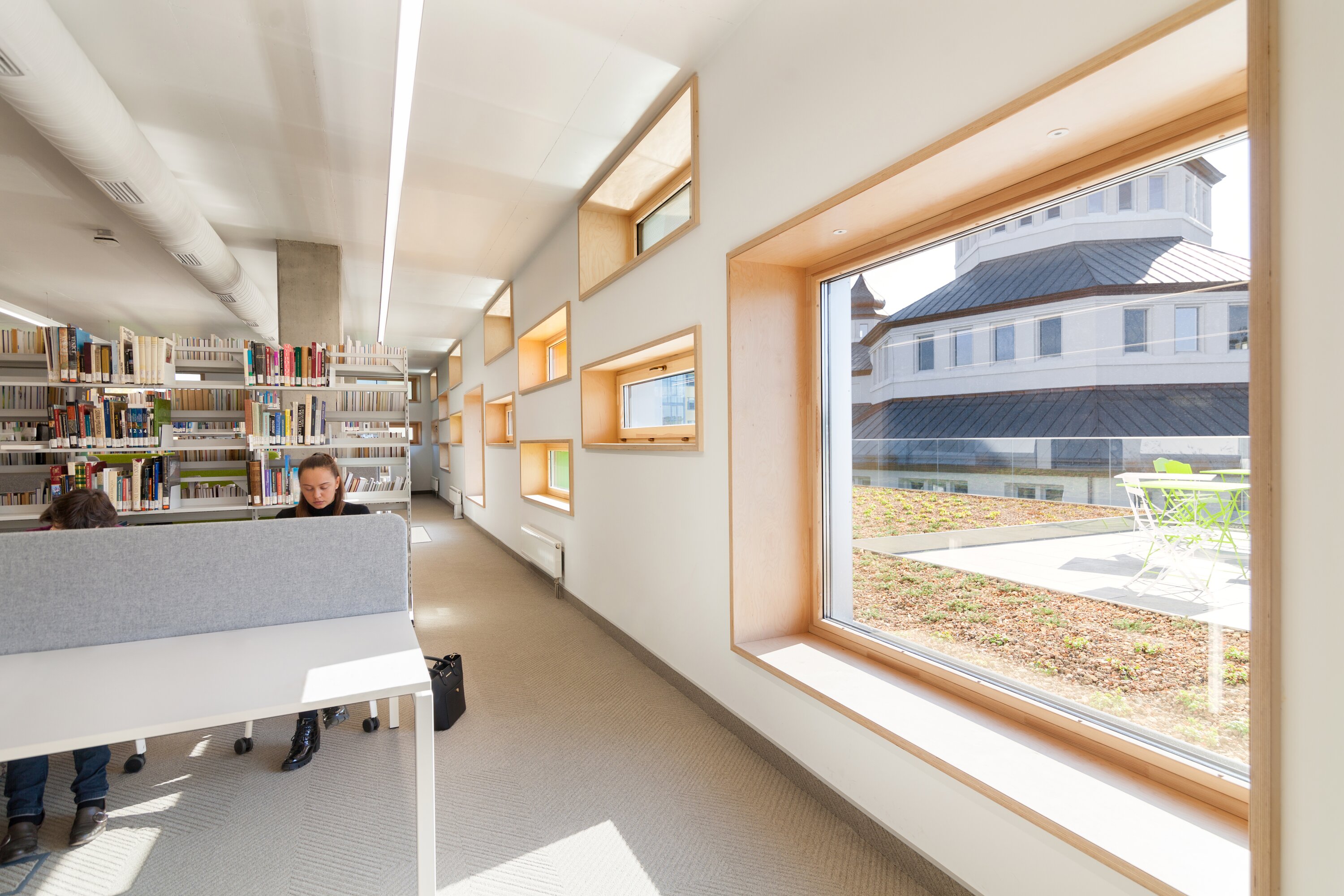 Project by Behnisch Architekturbuero, The Metropolitan Sheptytsky Center, Ukrainian Catholic University Lviv. Library study area with desks and bookshelves, small wood-framed windows. The view out of the window shows of a building and the terrace outside.