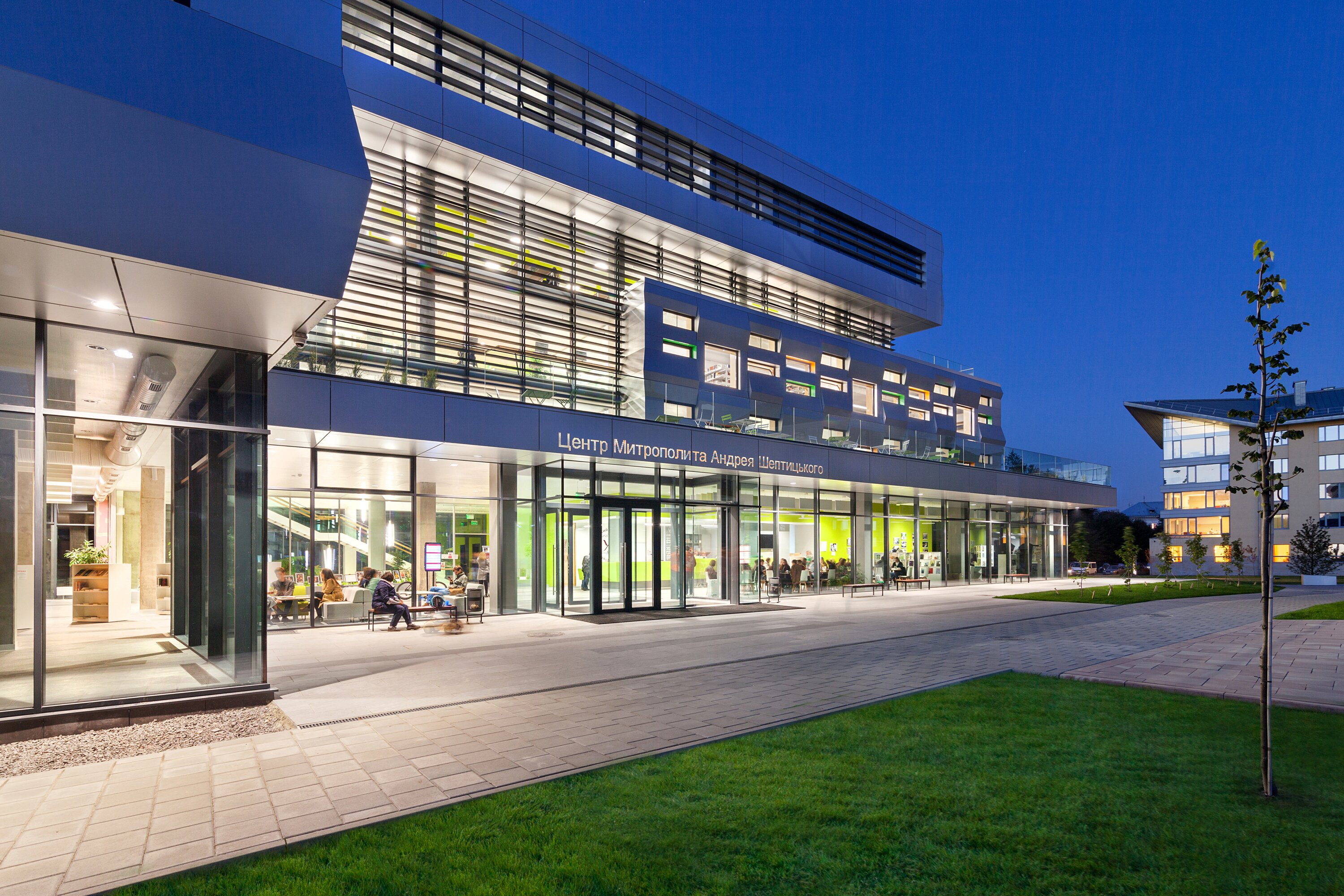 Project by Behnisch Architekturbuero, The Metropolitan Sheptytsky Center, Ukrainian Catholic University Lviv. The building facade with windows of varying sizes, offering a view into the library's interior. Tables and chairs are set up on a terrace in front.