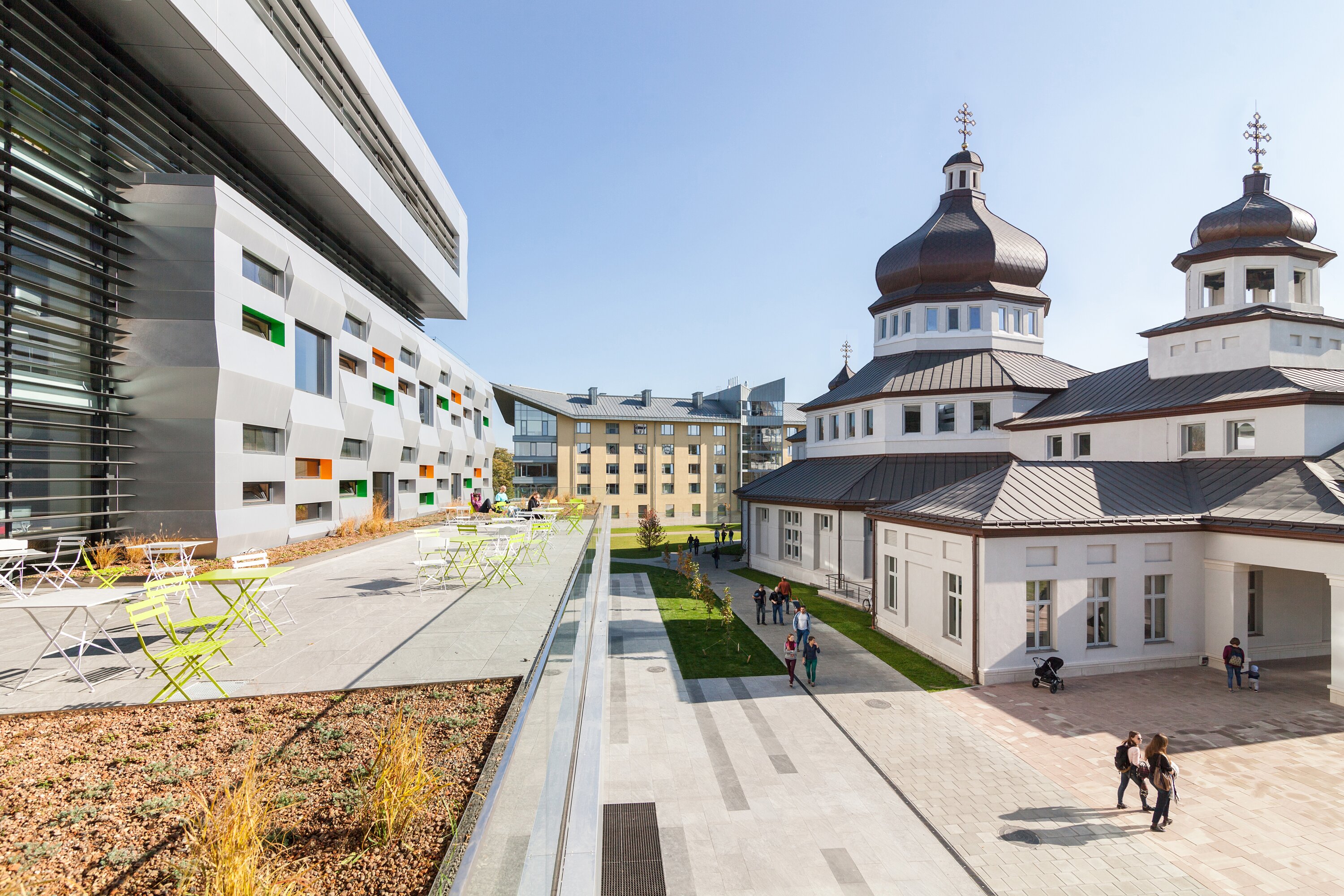 Project by Behnisch Architekturbuero, The Metropolitan Sheptytsky Center, Ukrainian Catholic University Lviv. Building with terrace seating beside a historic domed building, people walking along a landscaped path in a sunny courtyard.