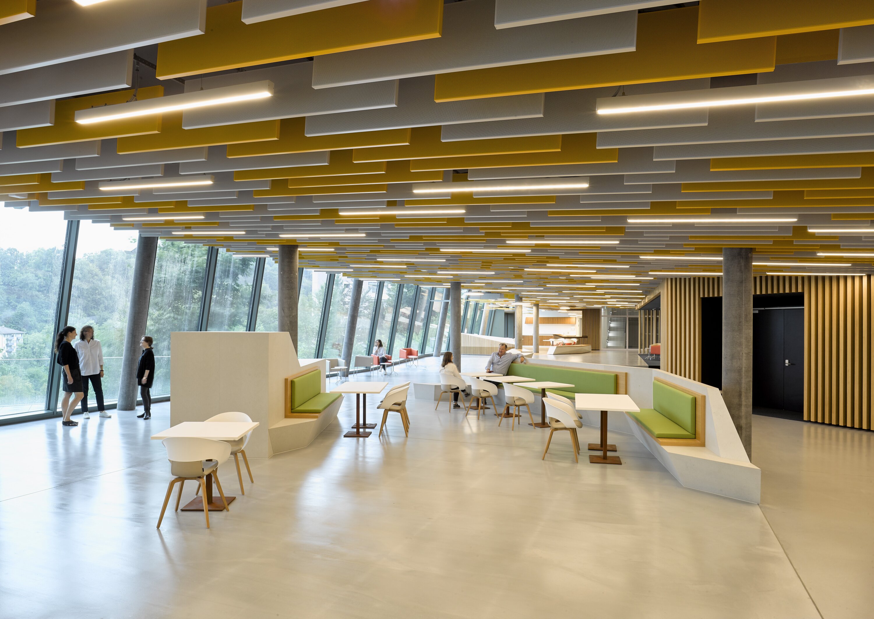 Project by Behnisch Architekturbuero, AGORA P&ocirc;le de recherche sur le cancer. Open-plan lounge with tables and seating. Large, slanted window facade and a ceiling made of yellow and white panels. Several people are sitting or standing in the room.