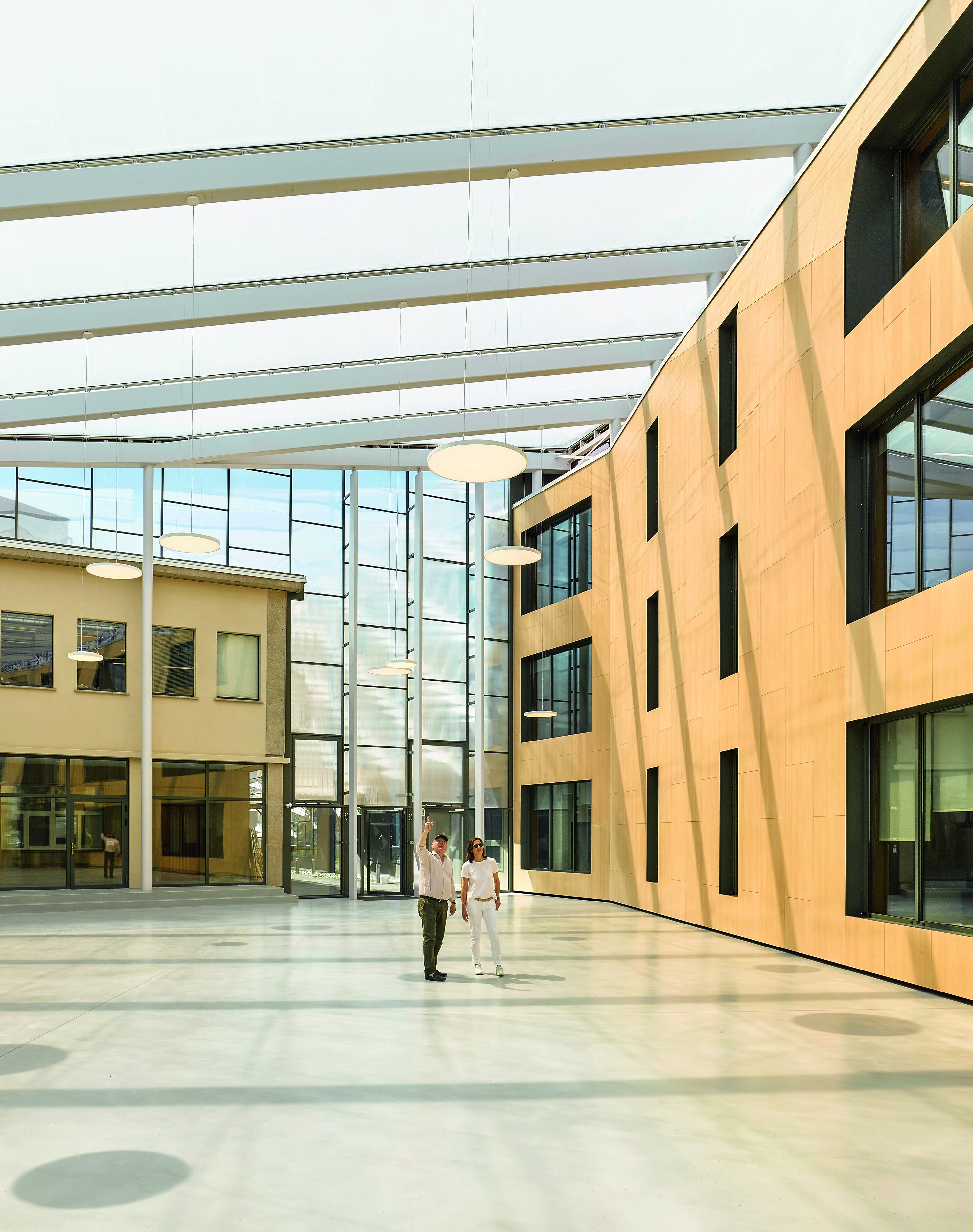 Project by Behnisch Architekturbuero, AGORA P&ocirc;le de recherche sur le cancer. A spacious atrium with a glass roof, light wood walls, and a glass facade. Two people are talking in the open interior.