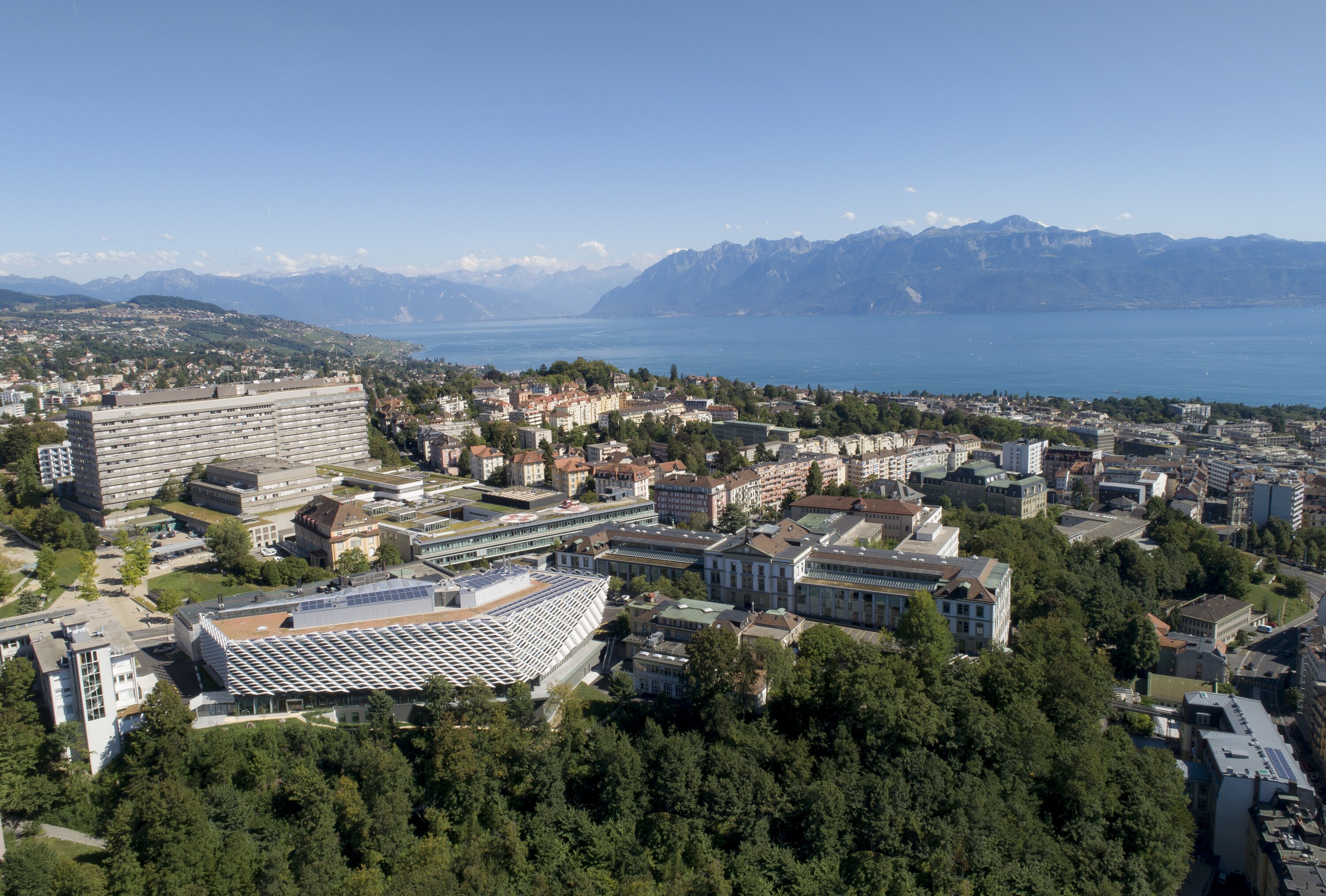 Project by Behnisch Architekturbuero, AGORA P&ocirc;le de recherche sur le cancer. Aerial view of a lakeside city with dense buildings. The large modern structure in the foreground, and mountains across the water under a clear sky.