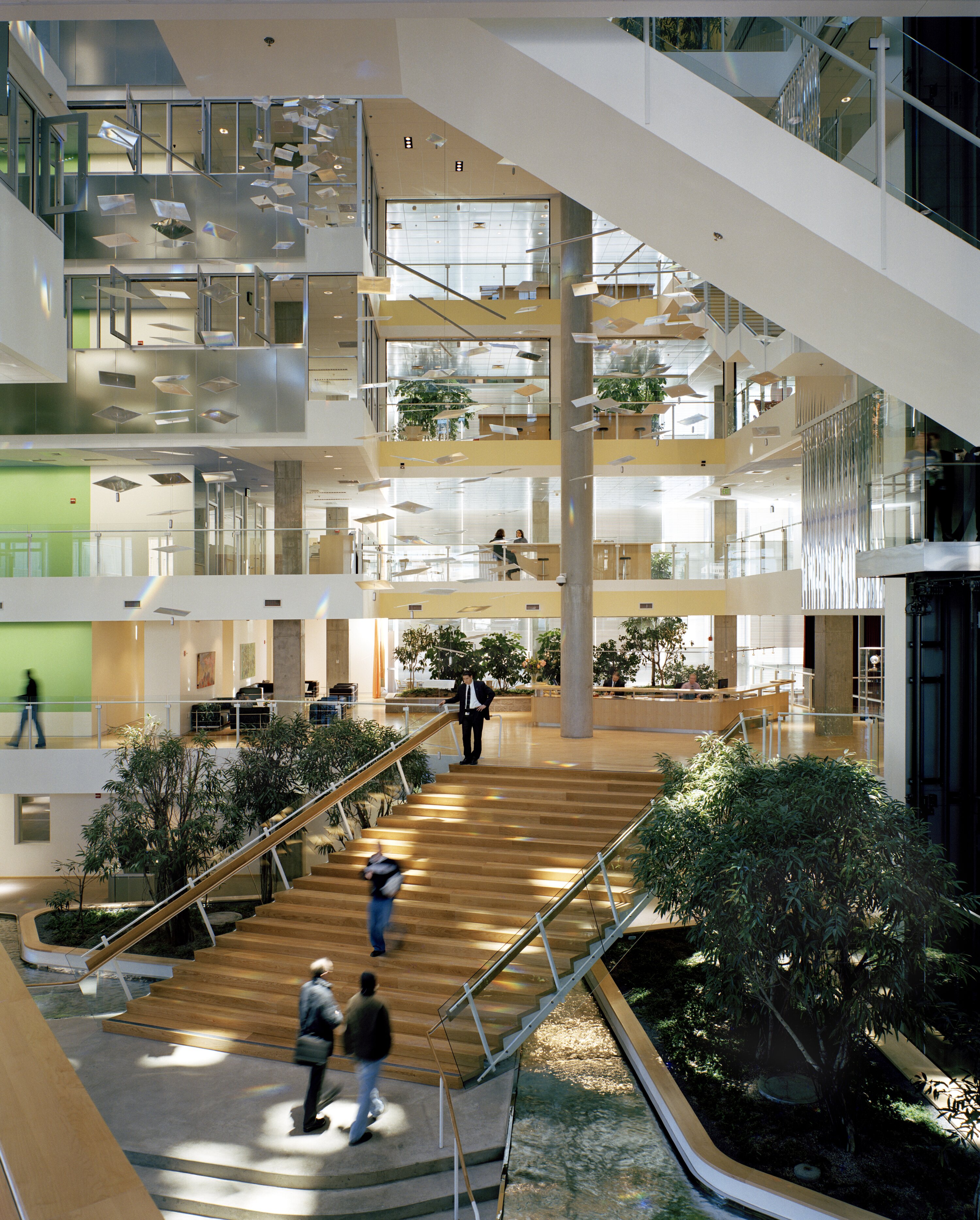 Project by Behnisch Architekturbuero, Genzyme Center. Large atrium with staircases, indoor trees, and multiple floors. People walk below a suspended installation of a floating sunlight redirection system.