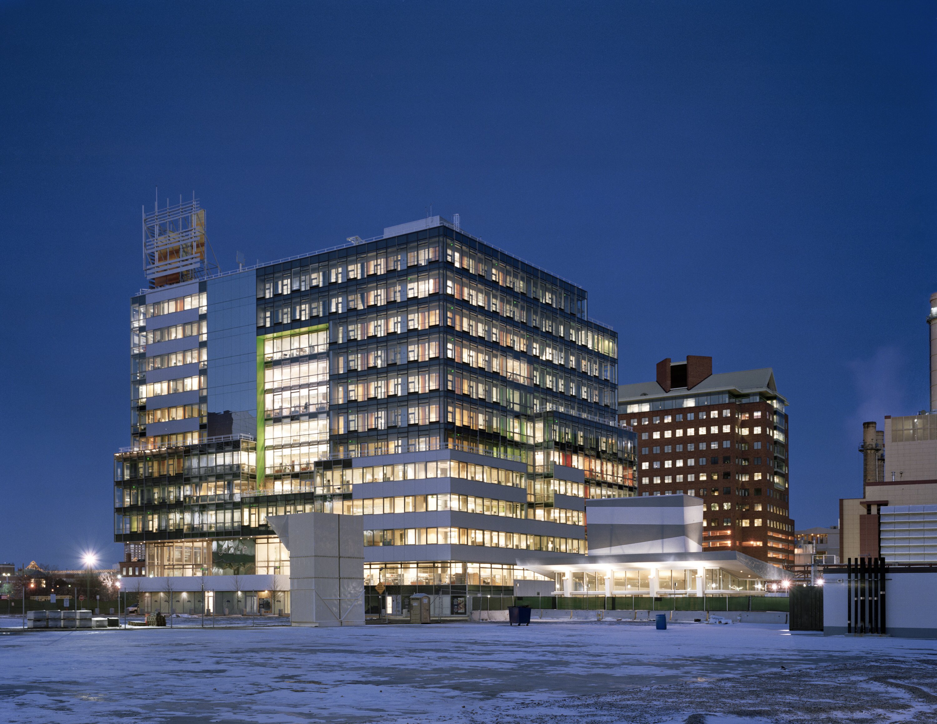 Project by Behnisch Architekturbuero, Genzyme Center. Illuminated modern office building at night with glass facade, surrounded by other buildings and a snowy open area in the foreground.