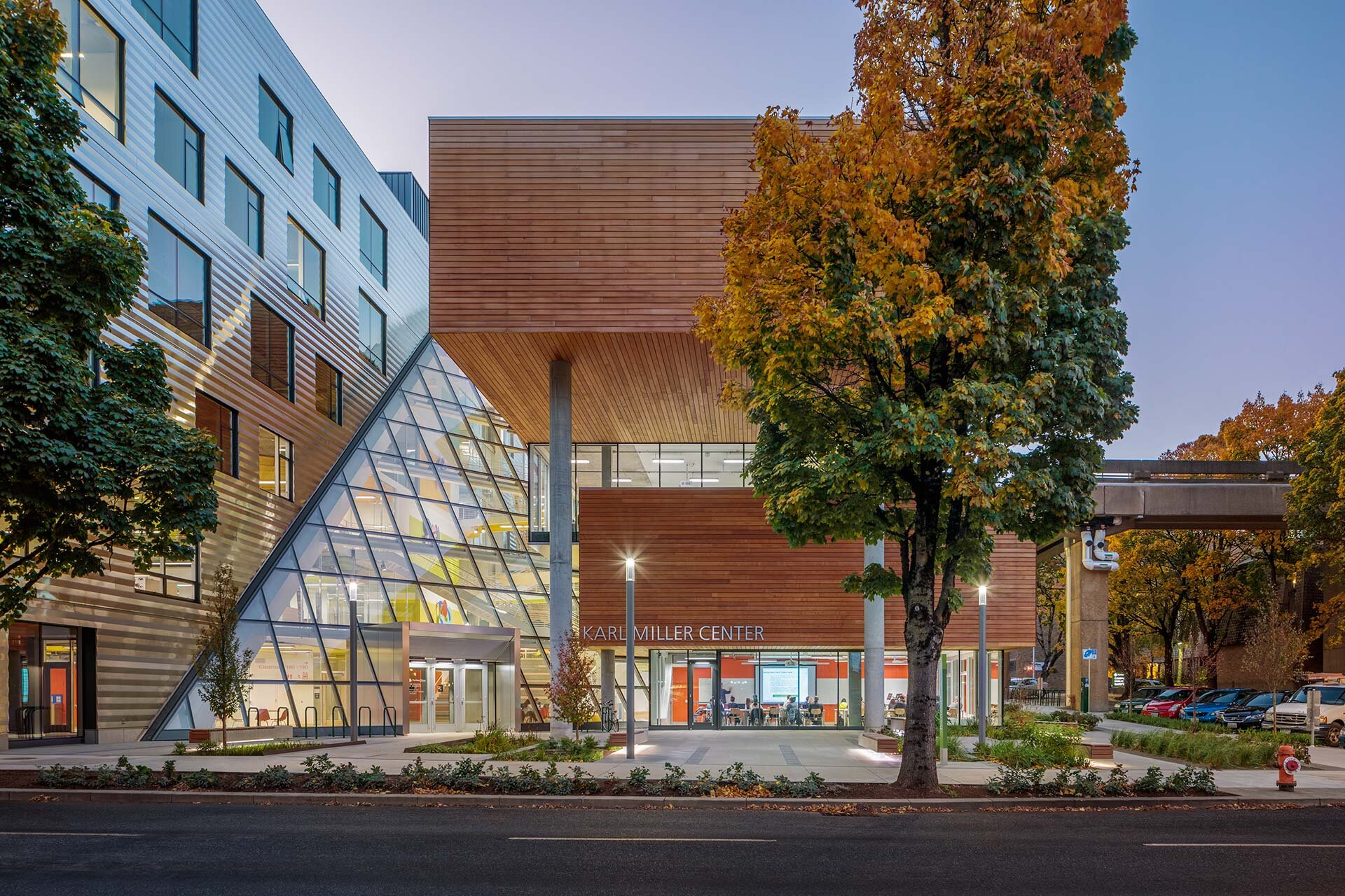 Project by Behnisch Architekturbuero, Karl Miller Center. Campus building with glass pyramid entrance and wood facade. Trees in autumn, and people visible inside at dusk.