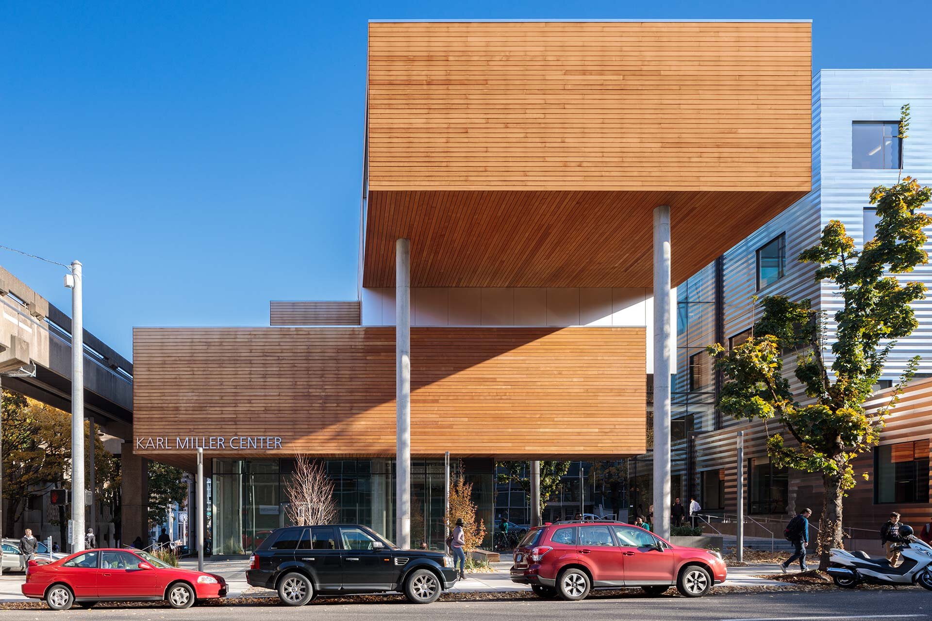 Project by Behnisch Architekturbuero, Karl Miller Center. Street view of the Karl Miller Center, showing a large, wood-clad building on stilts with a glass entrance below. Cars are parked in front of the building along the street.