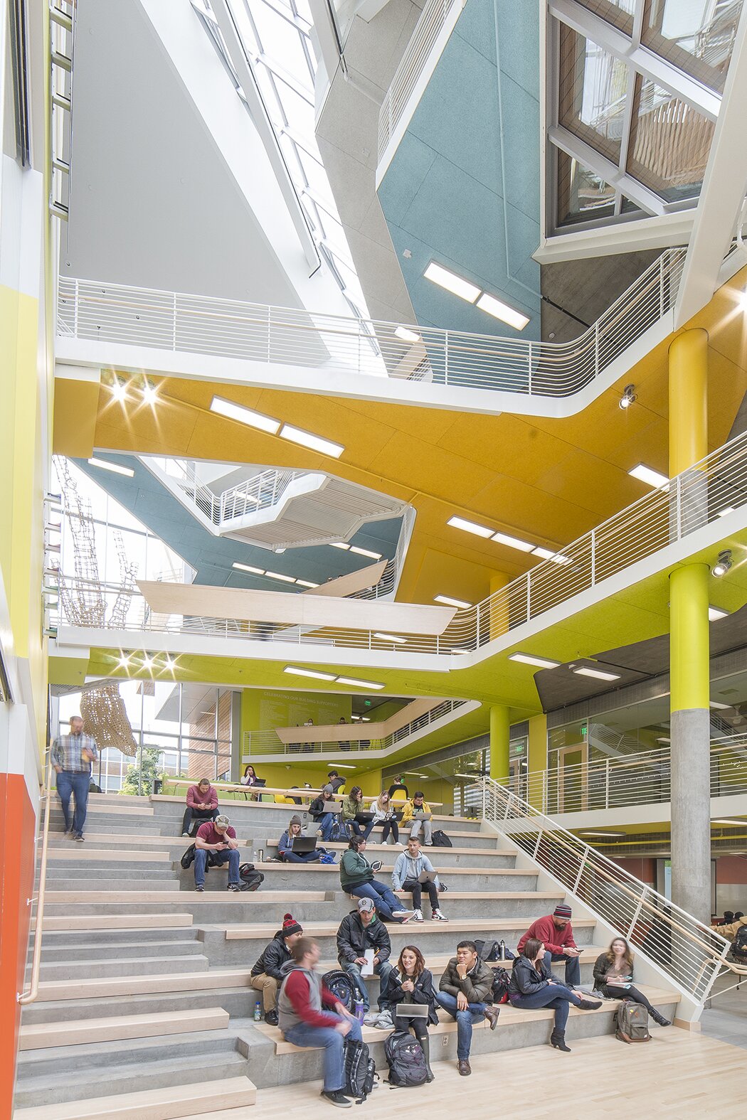 Project by Behnisch Architekturbuero, Karl Miller Center. A large atrium with terraced seating. Students sit and chat on the steps. The ceilings are brightly colored.