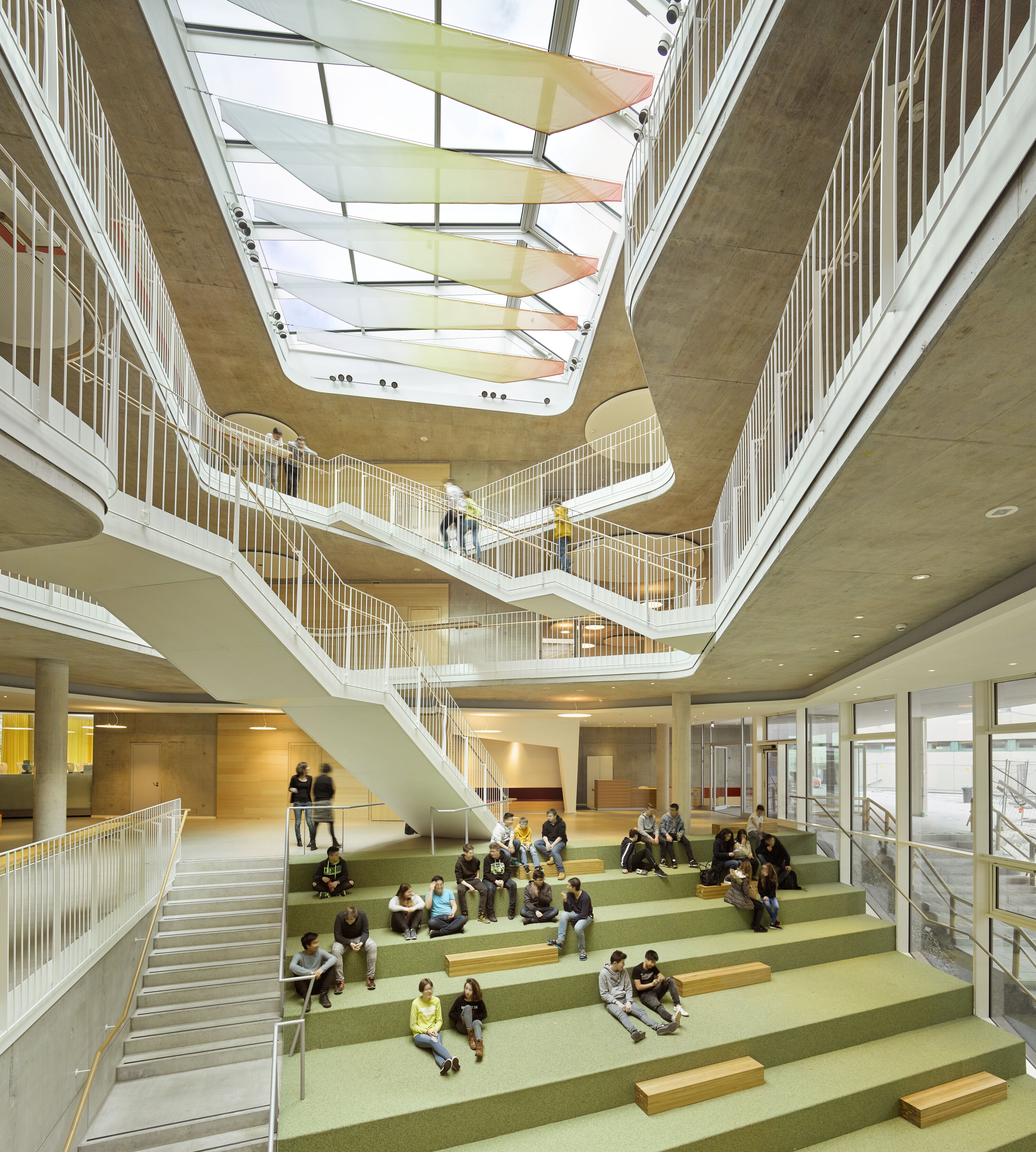 Project by Behnisch Architekturbuero, Anna-Pr&ouml;ll Secondary School and Sports Hall. Large school atrium with tiered seating, students sitting and talking, multiple staircases and walkways under a skylight ceiling.