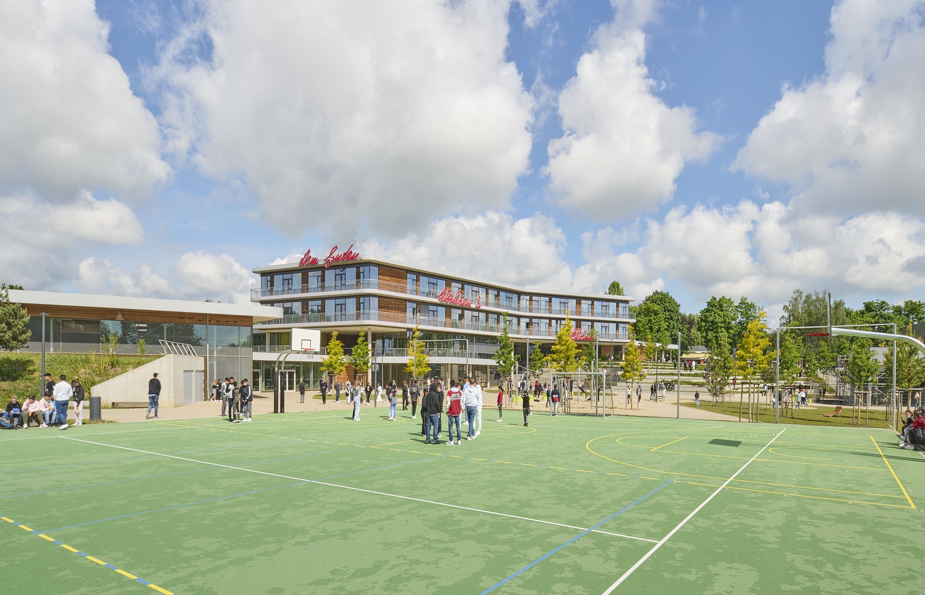 Project by Behnisch Architekturbuero, Anna-Pr&ouml;ll Secondary School and Sports Hall. School campus with students gathered on an outdoor sports court. In the background the Schoolbuilding, trees and a playground equipment nearby.