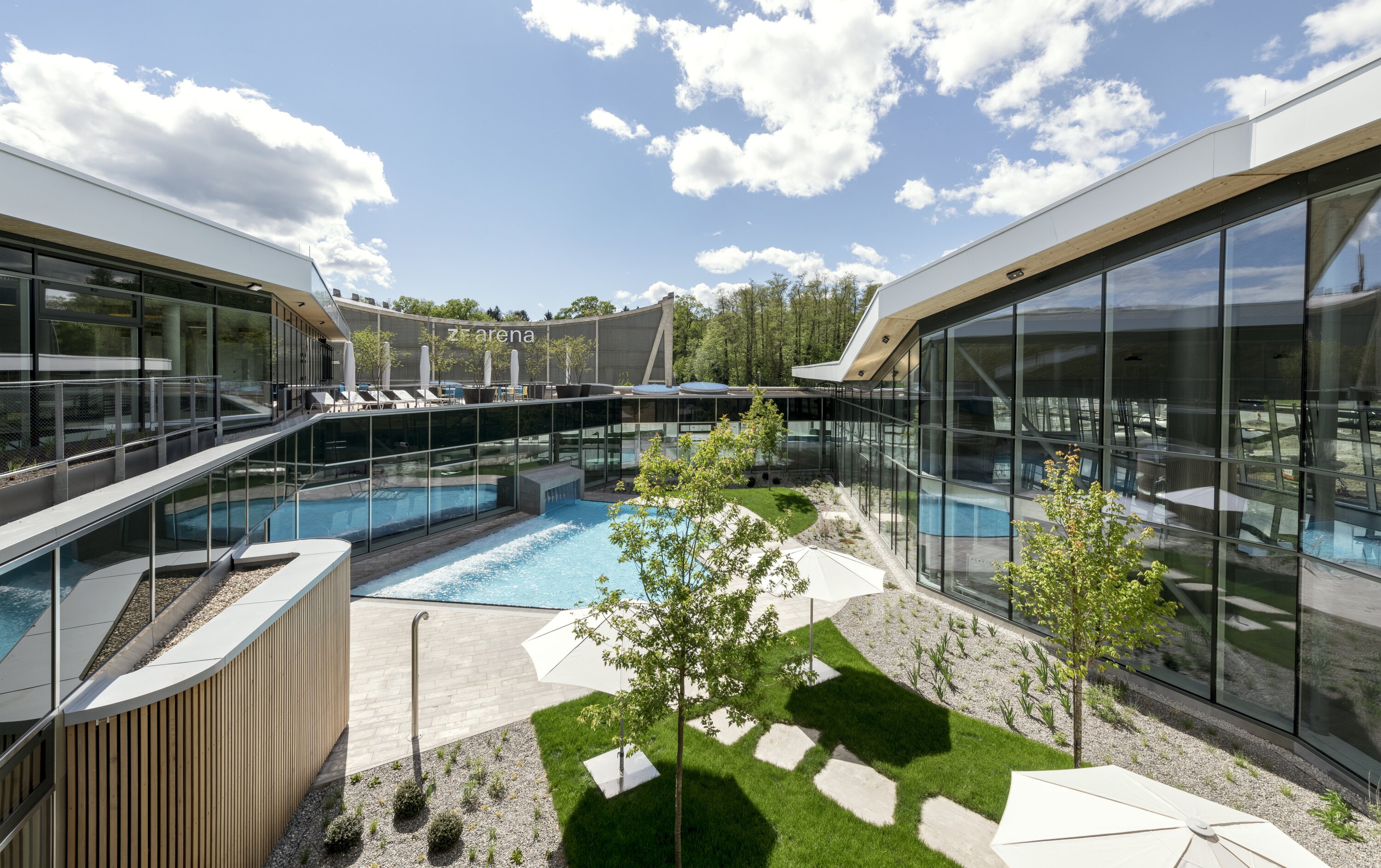 Project by Behnisch Architekturbuero, Sportbad Friedrichshafen. Courtyard with glass buildings around a pool, landscaped garden with trees and umbrellas, and terraces under a partly cloudy sky.