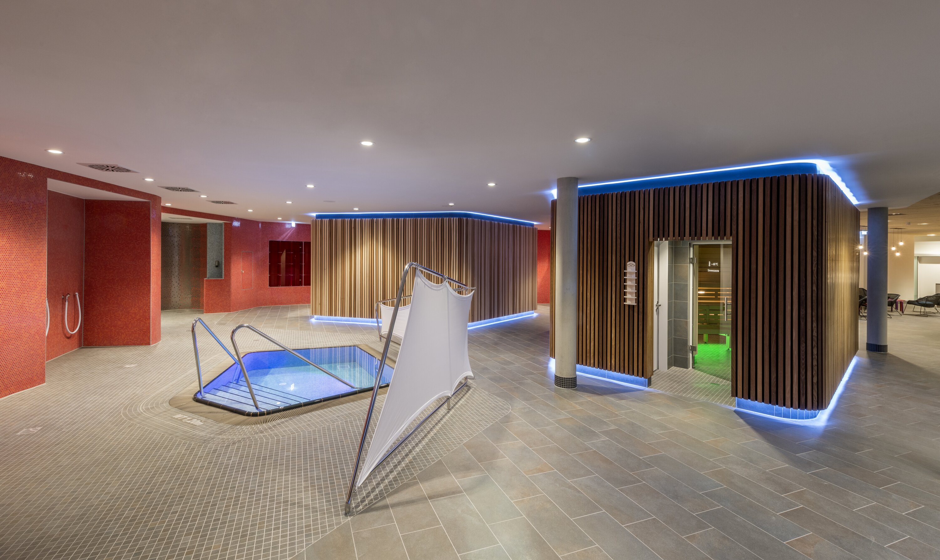 Project by Behnisch Architekturbuero, Sportbad Friedrichshafen. Indoor wellness area with a small plunge pool, tiled floor, and wood-paneled sauna rooms. The left side of the room features red walls, and the sauna rooms are surrounded by blue accent lighting.