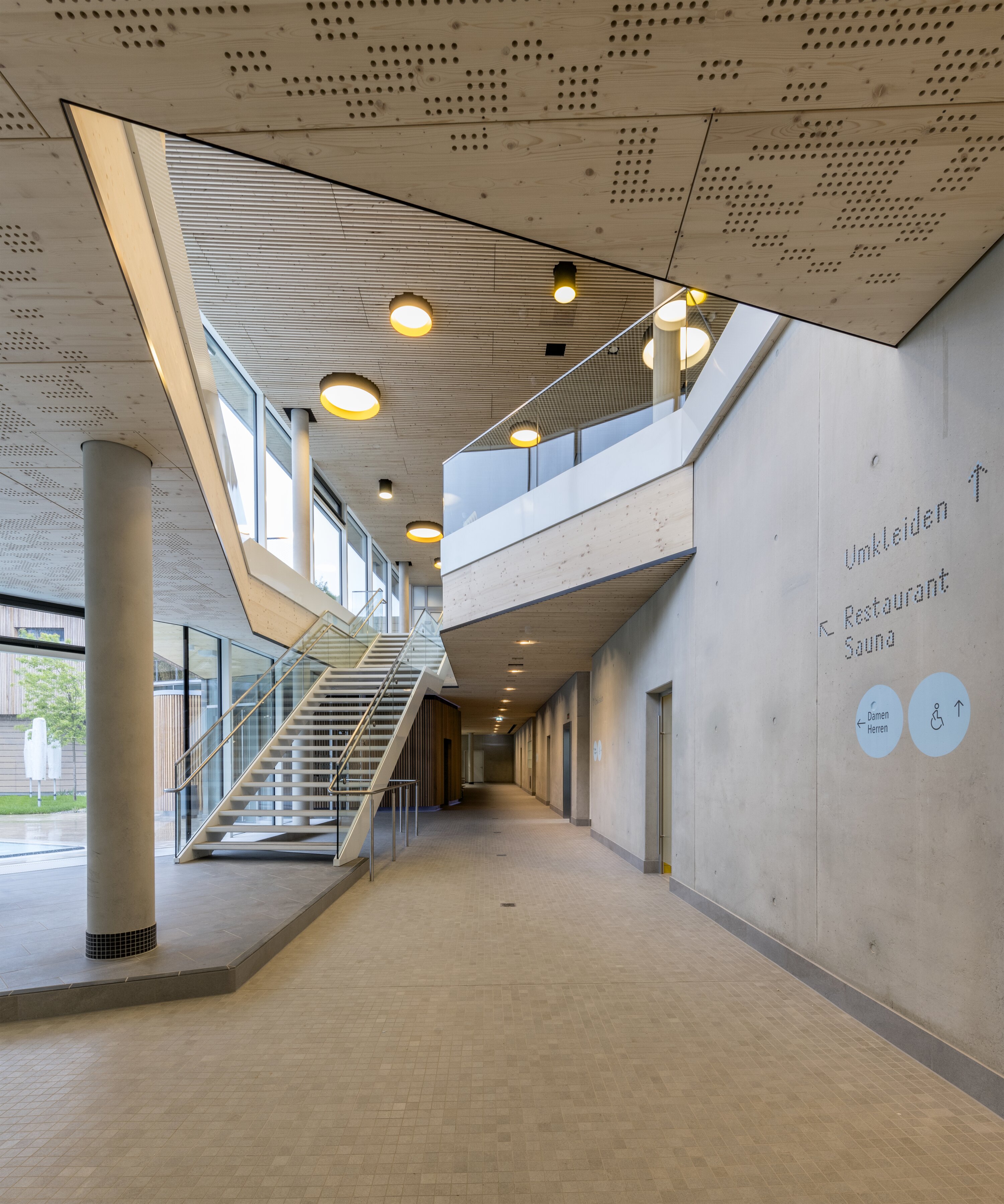 Project by Behnisch Architekturbuero, Sportbad Friedrichshafen. Modern interior corridor with staircase, glass railing, wood ceiling panels, concrete walls, and directional signage for changing rooms and sauna.