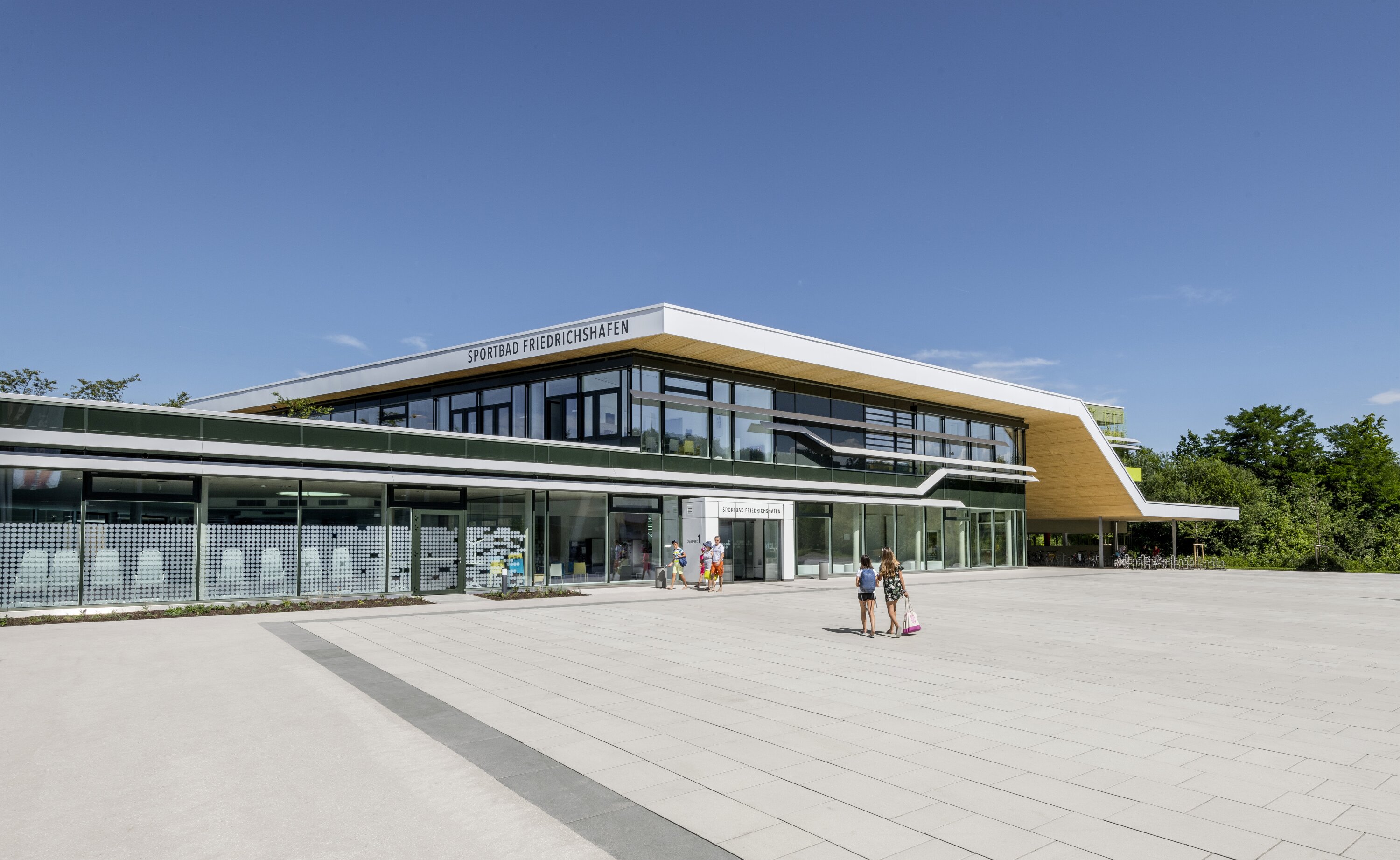 Project by Behnisch Architekturbuero, Sportbad Friedrichshafen. A modern sports pool with a glass facade and cantilevered roof; people strolling across a large paved square under a clear blue sky.