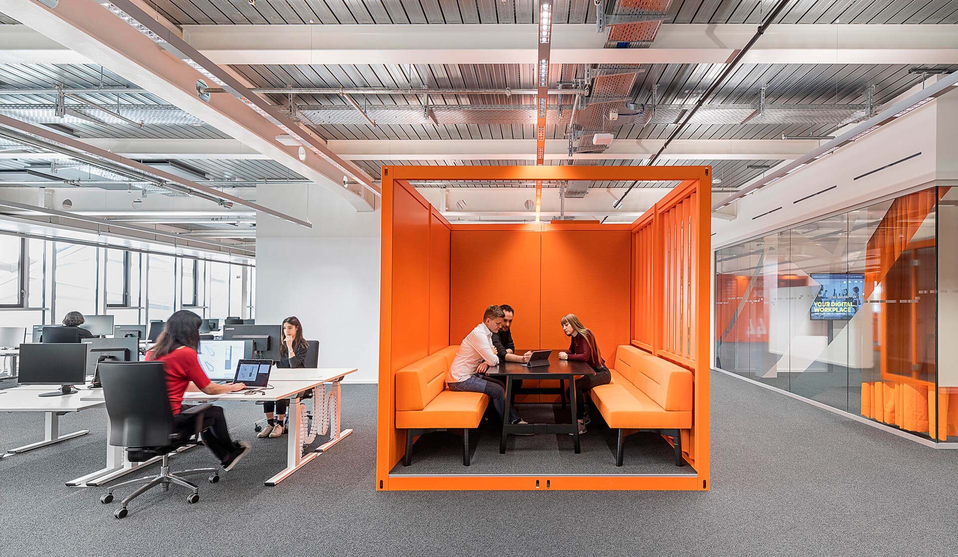 Project by Behnisch Architekturbuero, adidas ARENA. Open-plan office with desks and computers, and an orange meeting booth where three people sit around a table working together.