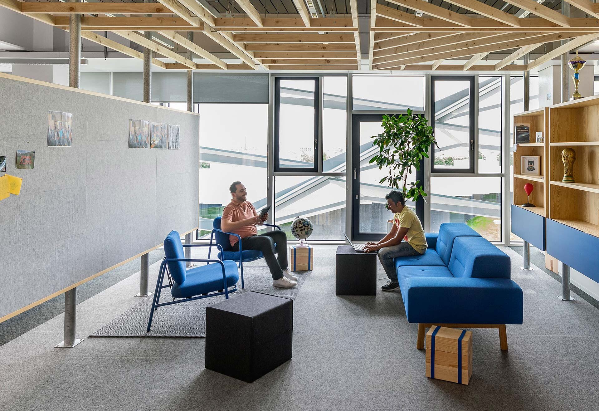 Project by Behnisch Architekturbuero, adidas ARENA. Lounge area with blue seating and small tables, two people working on devices, large windows in the background, indoor plant, and shelving with objects.
