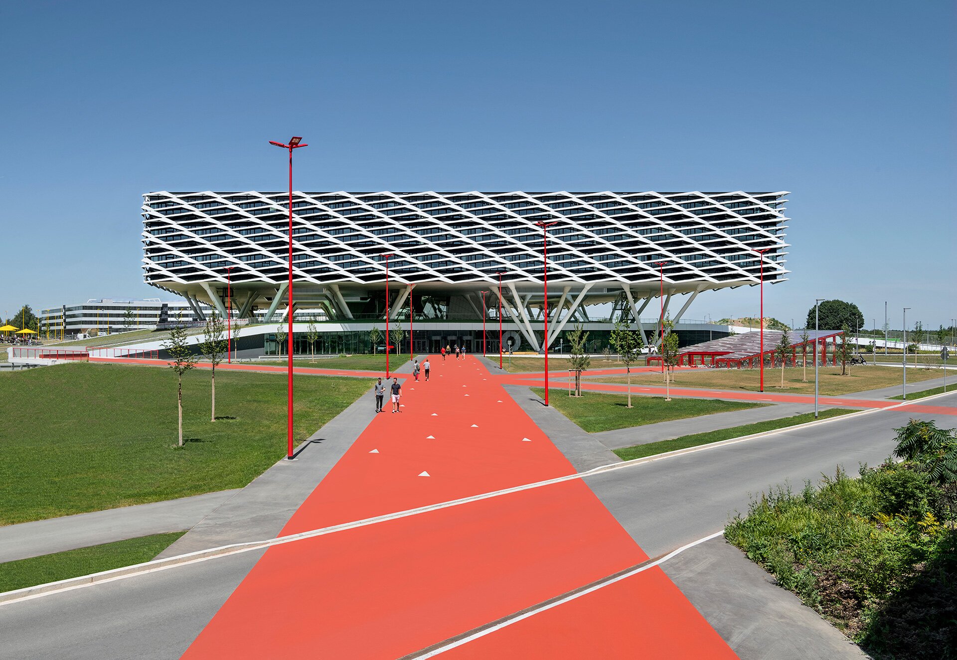 Project by Behnisch Architekturbuero, adidas ARENA.  A large, modern building with a sloping, grid-like facade features it is a specially designed sun protection and shading system. The main building is elevated and stands on stilts. People walk along a wide red path leading to the entrance.