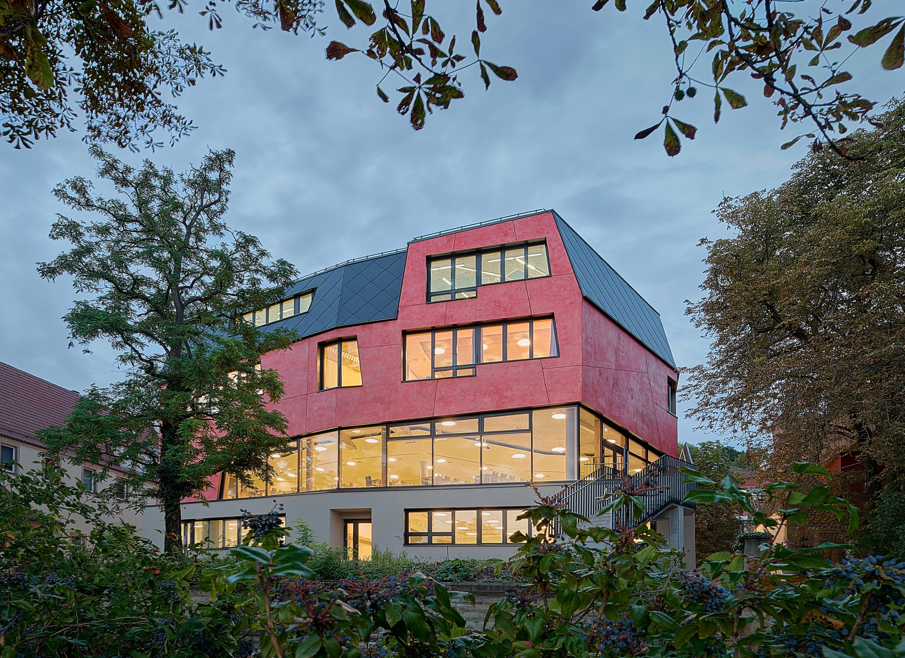 Project by Behnisch Architekturbuero, Freie Waldorfschule Uhlandshoehe - New School and Administration Building. Red multi-story building with large lit windows, surrounded by trees, viewed at dusk.