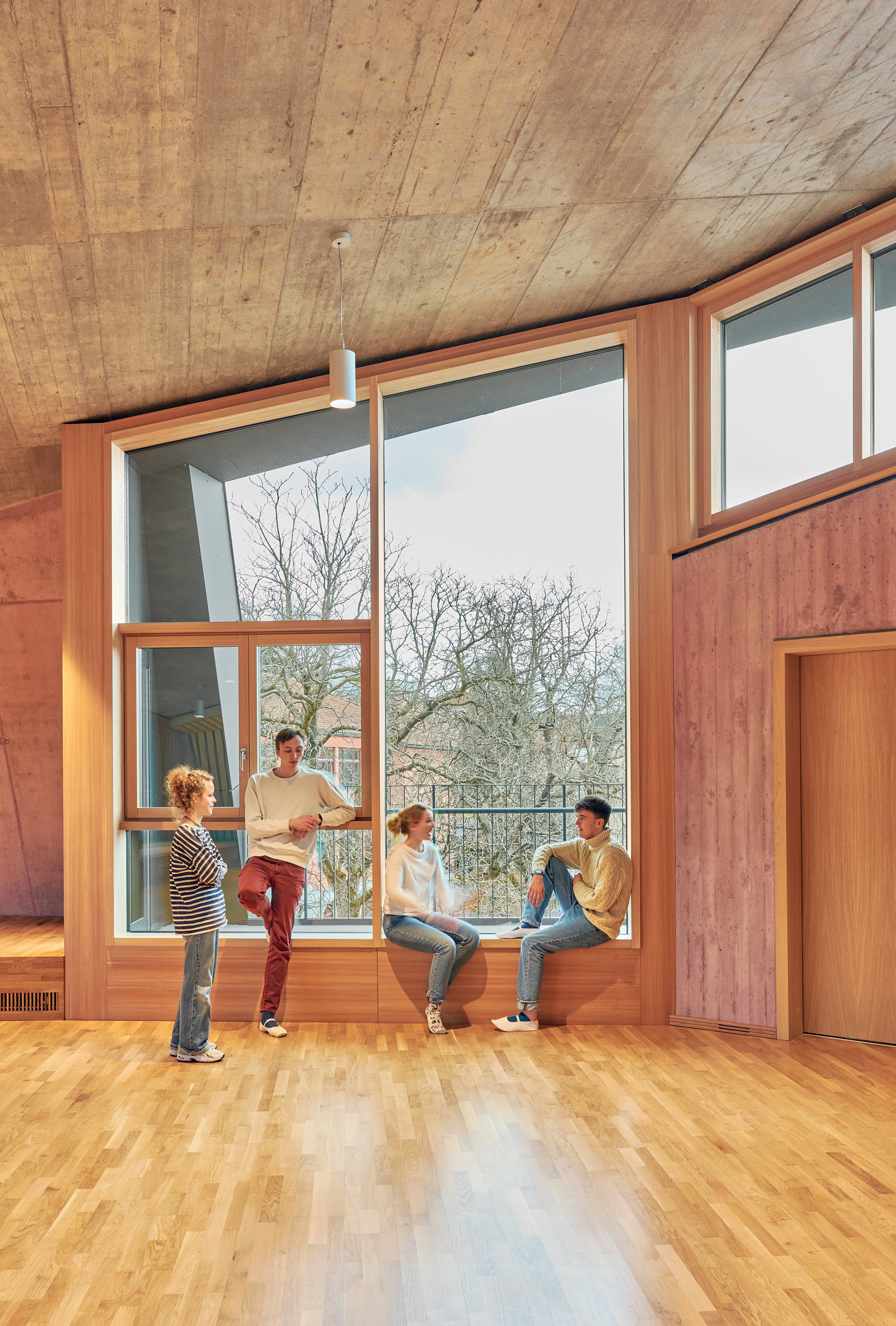 Project by Behnisch Architekturbuero, Freie Waldorfschule Uhlandshoehe - New School and Administration Building. Four people talking by large window in wood interior room, two seated on window ledge and two standing nearby.
