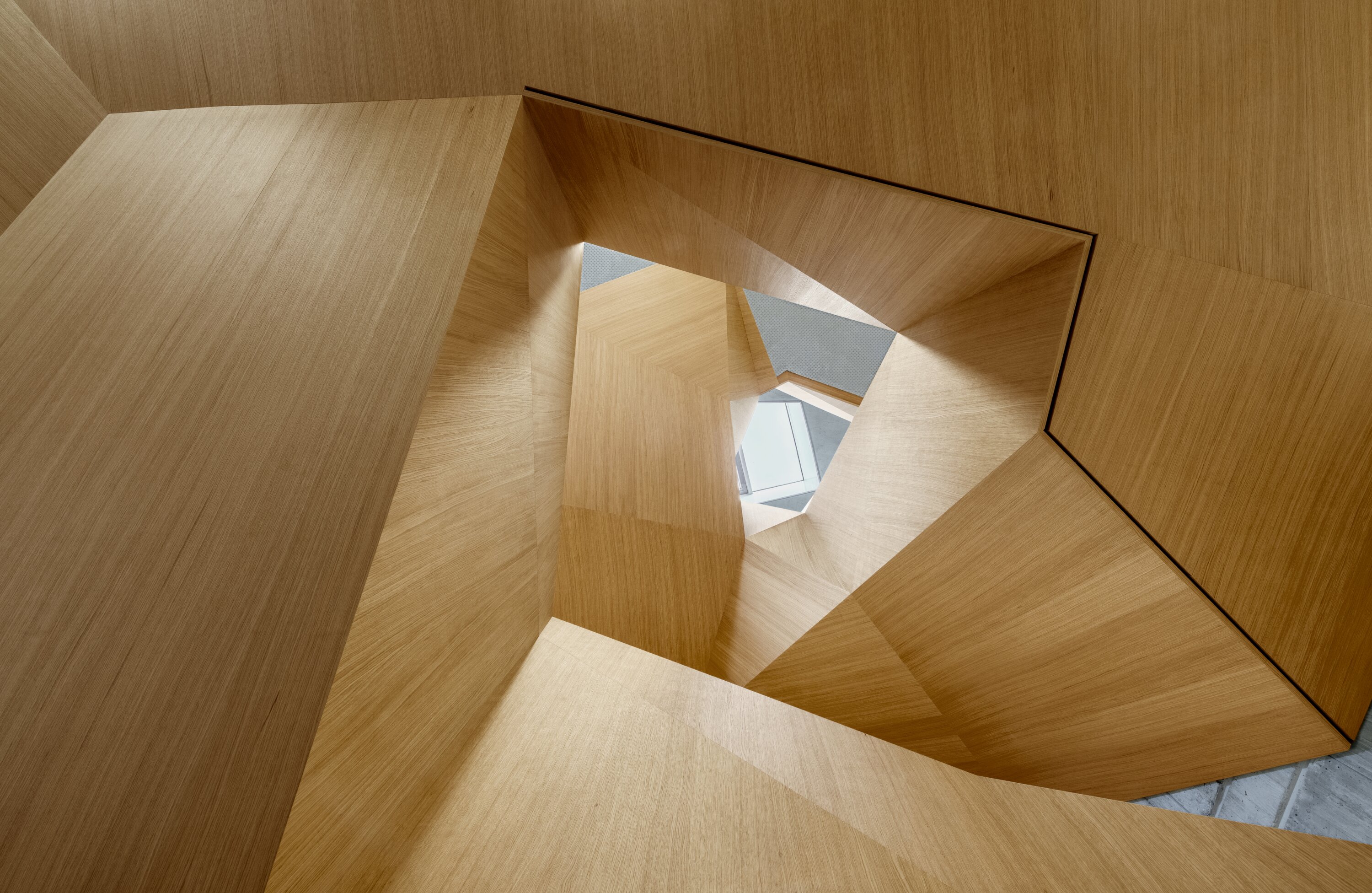 Project by Behnisch Architekturbuero, Freie Waldorfschule Uhlandshoehe - New School and Administration Building. View upward through angular wooden stairwell with faceted walls and skylight opening above