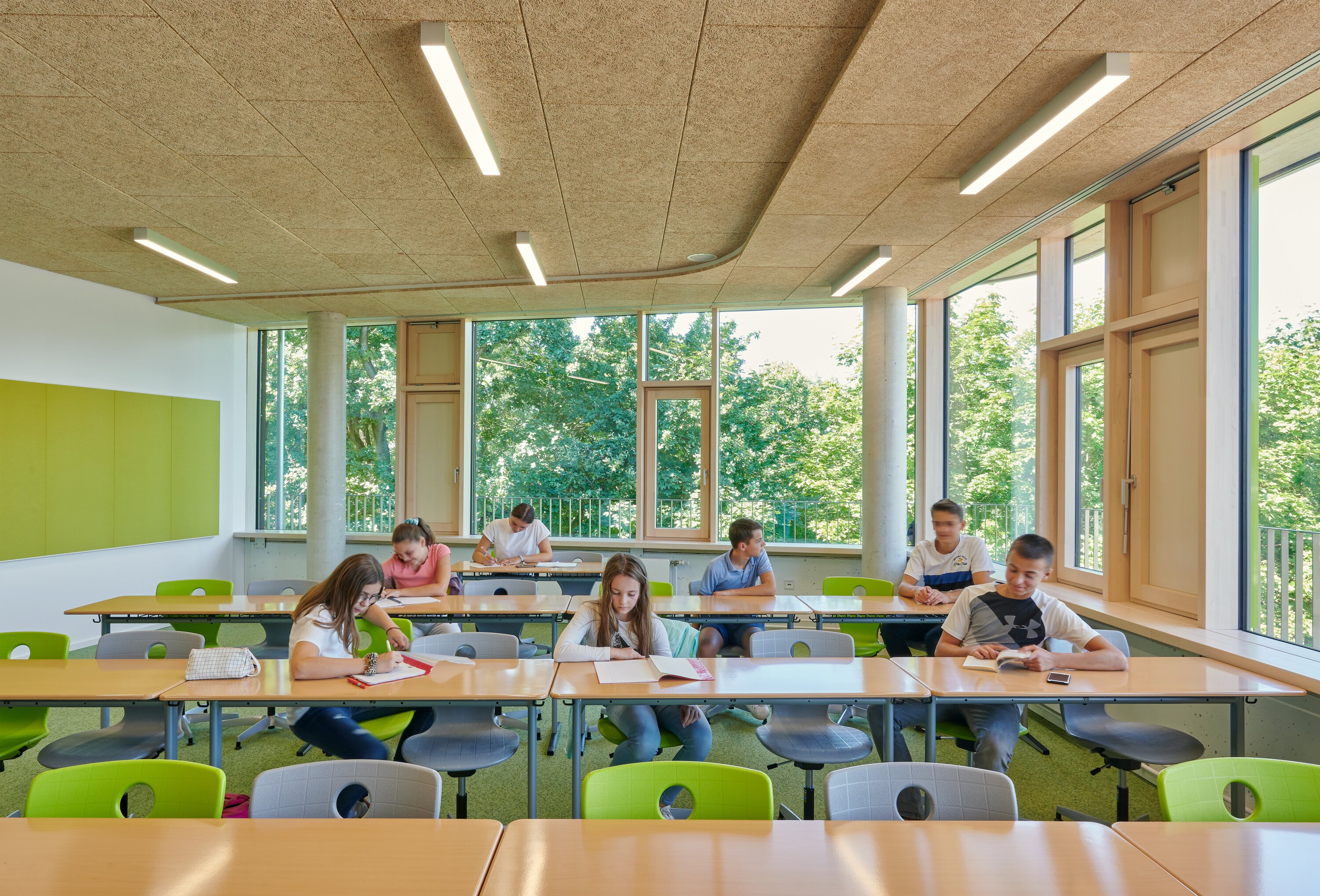 Project by Behnisch Architekturbuero, Lurup Neighborhood School. A classroom with students reading and writing. Large windows overlooking trees outside; the room is furnished with tables, green chairs, and a cork ceiling.