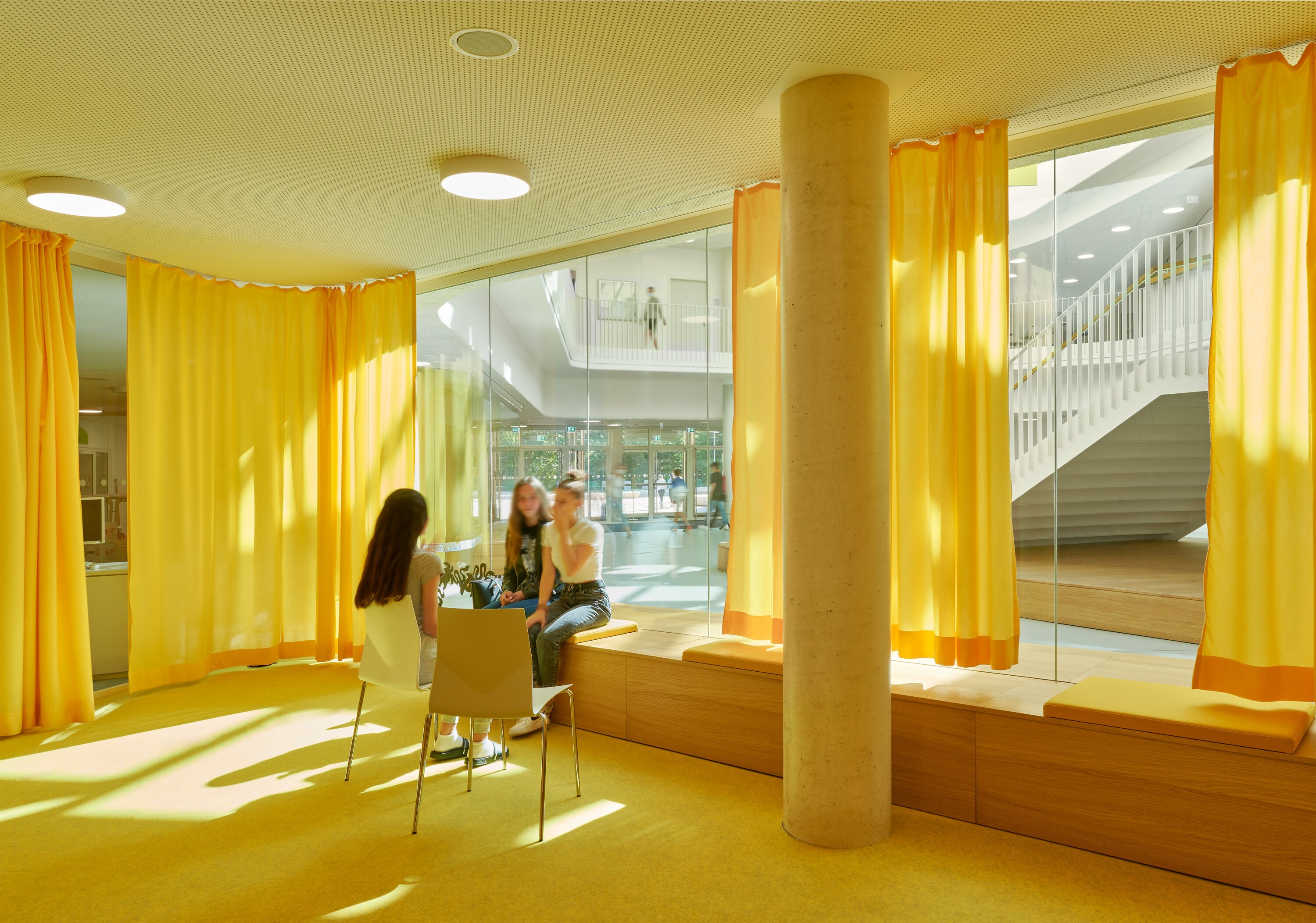 Project by Behnisch Architekturbuero, Lurup Neighborhood School. Small lounge with yellow curtains and carpet, students sitting and talking near a window, with view into adjacent atrium and stairs.