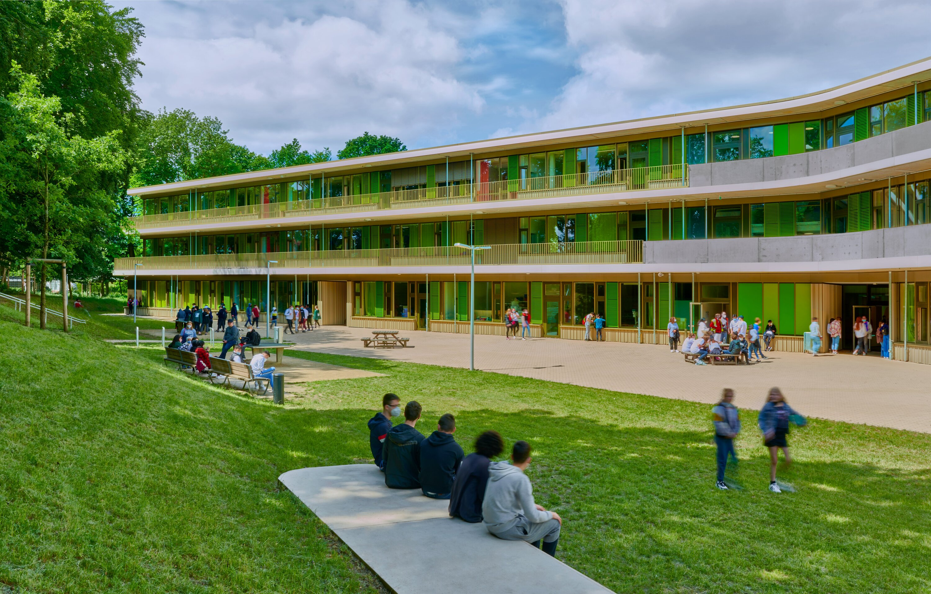 Project by Behnisch Architekturbuero, Lurup Neighborhood School. School building with green facade and balconies, students have a break in the courtyard and sitting on benches under the trees.