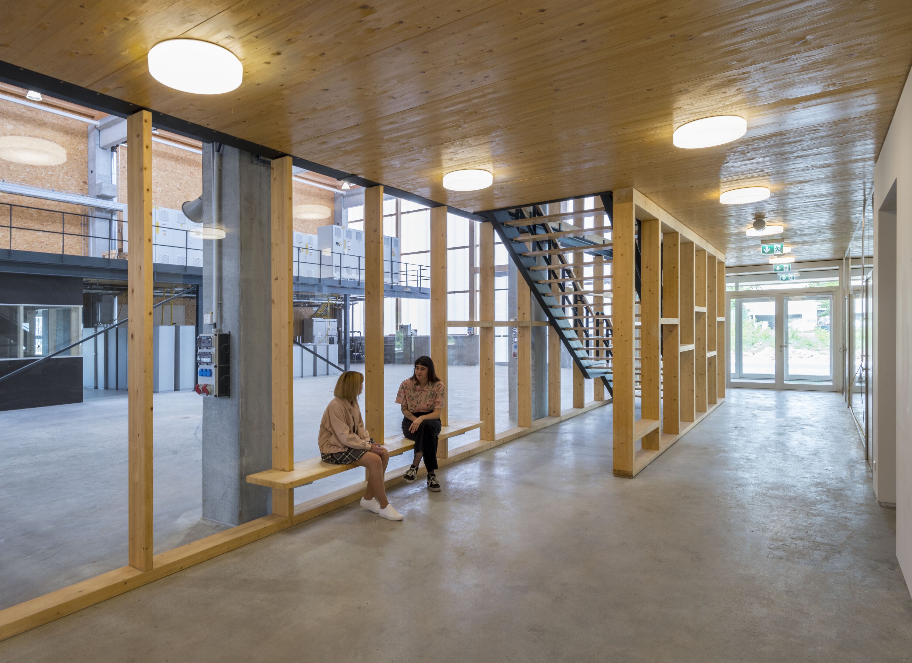Project by Behnisch Architekturbuero, KIT Energy Lab 2.0. Entrance area and hallway with a wooden roof structure, skylights, and exposed beams. Light-filled space thanks to an open glass facade. Two women sit on a wooden bench and talk.