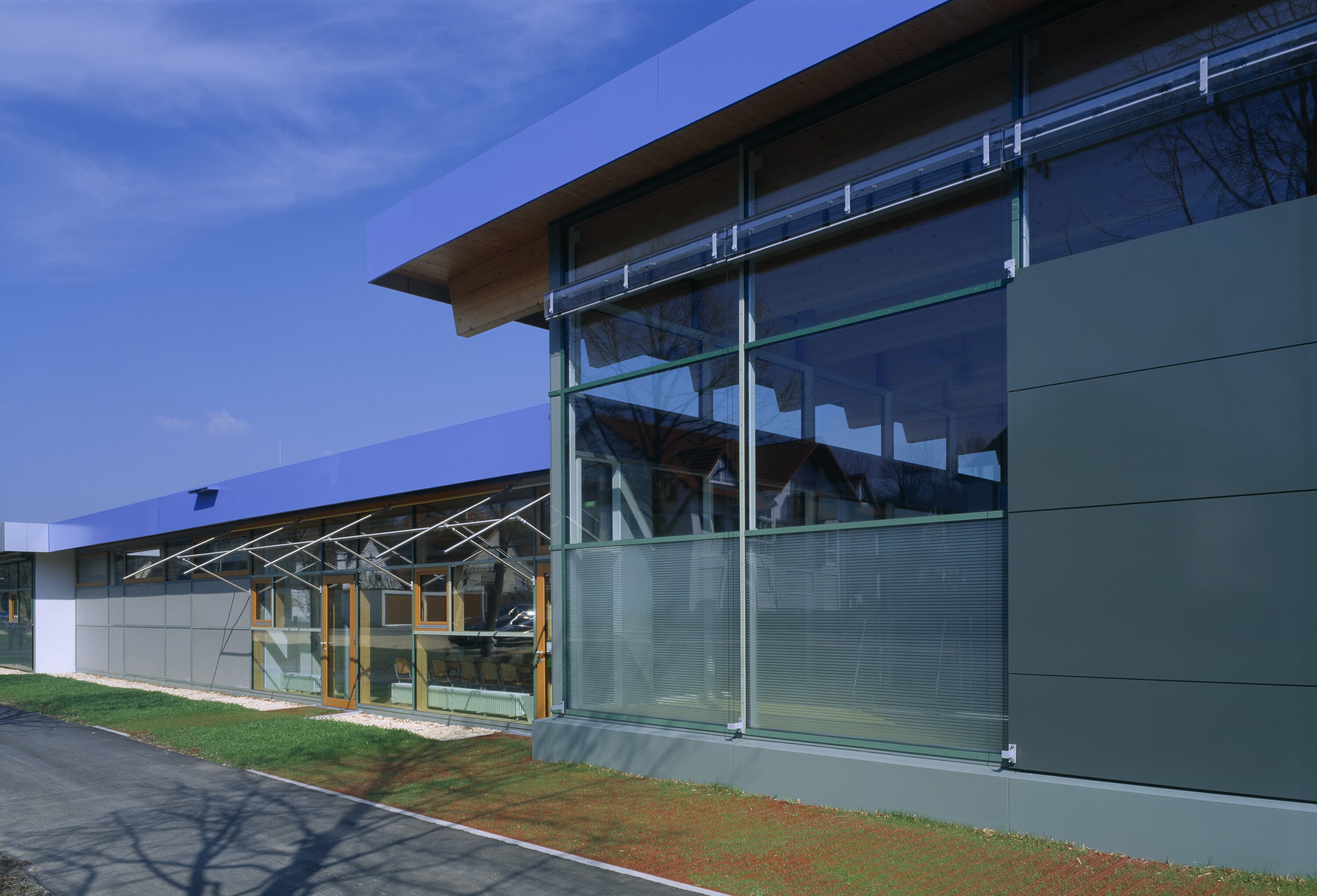 Project by Behnisch Architekturbuero, Pistorius School Herbrechtingen. Modern school building with glass facade, closed panels and wooden elements next to a walkway and lawn under a blue sky.