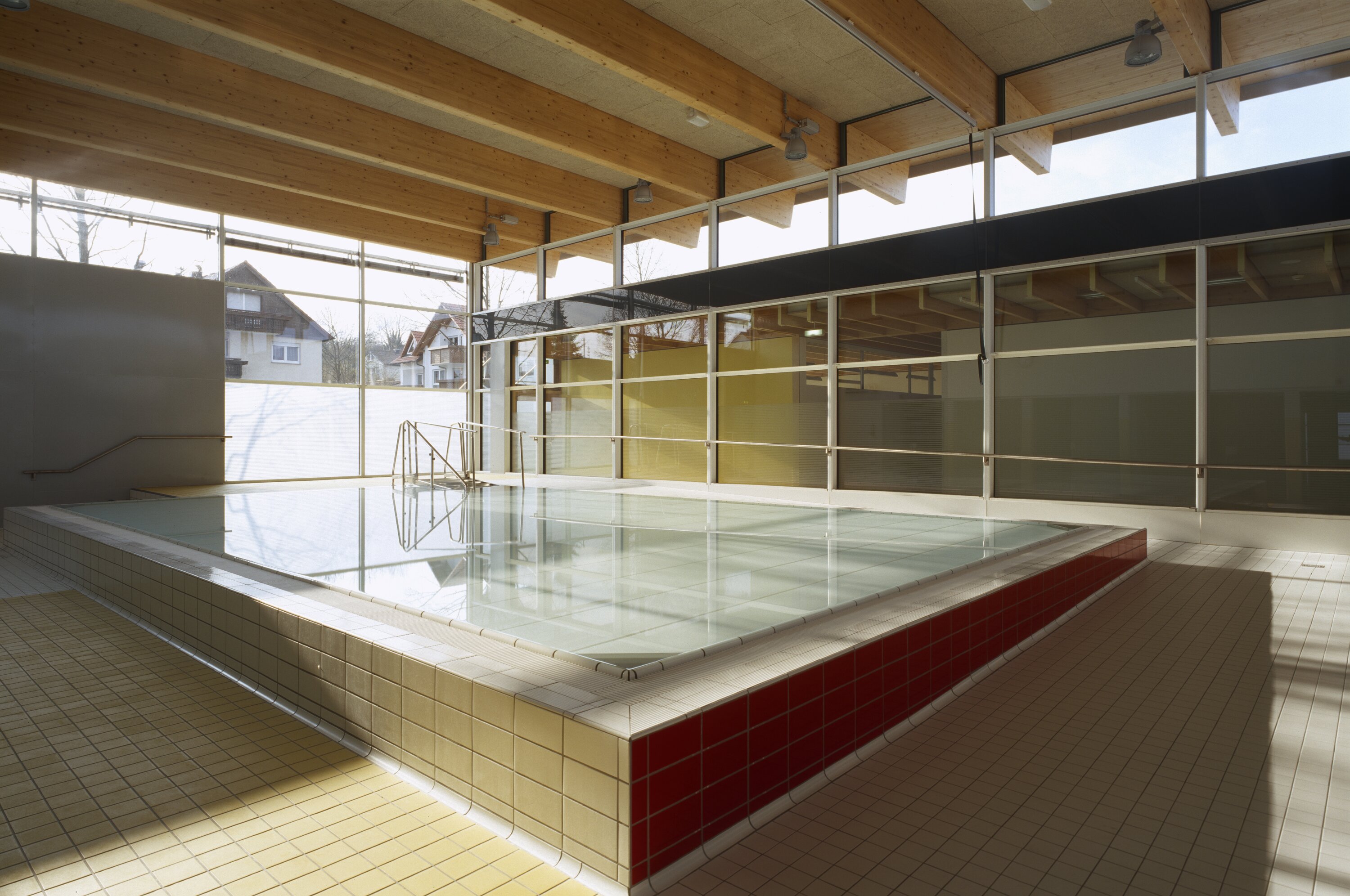 Project by Behnisch Architekturbuero, Pistorius School Herbrechtingen. Indoor pool with tiled edges and metal handrails, set in a sunlit room with large windows and a wood-beamed ceiling.