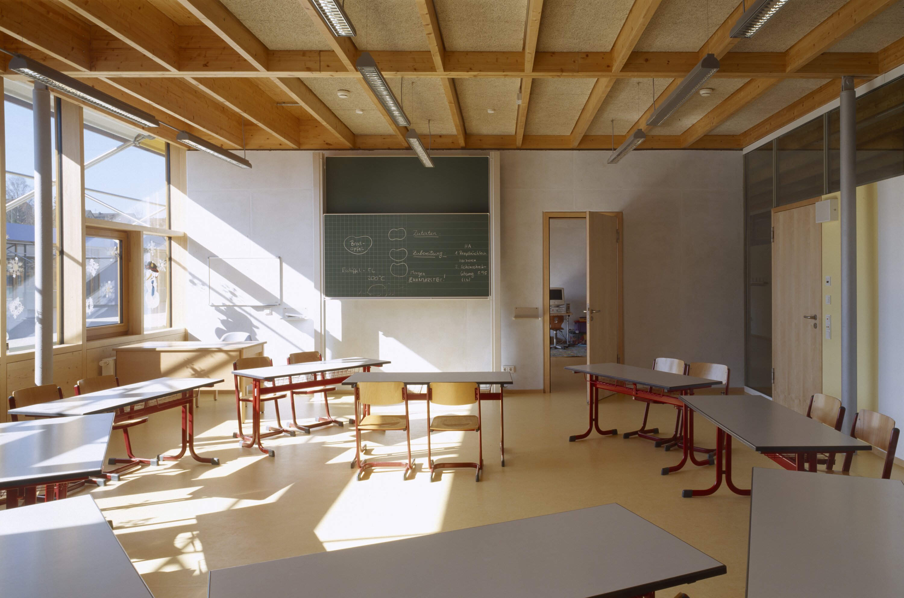 Project by Behnisch Architekturbuero, Pistorius School Herbrechtingen. Classroom with desks and chairs, chalkboard on wall, large windows letting in sunlight, and a wood-beamed ceiling overhead.