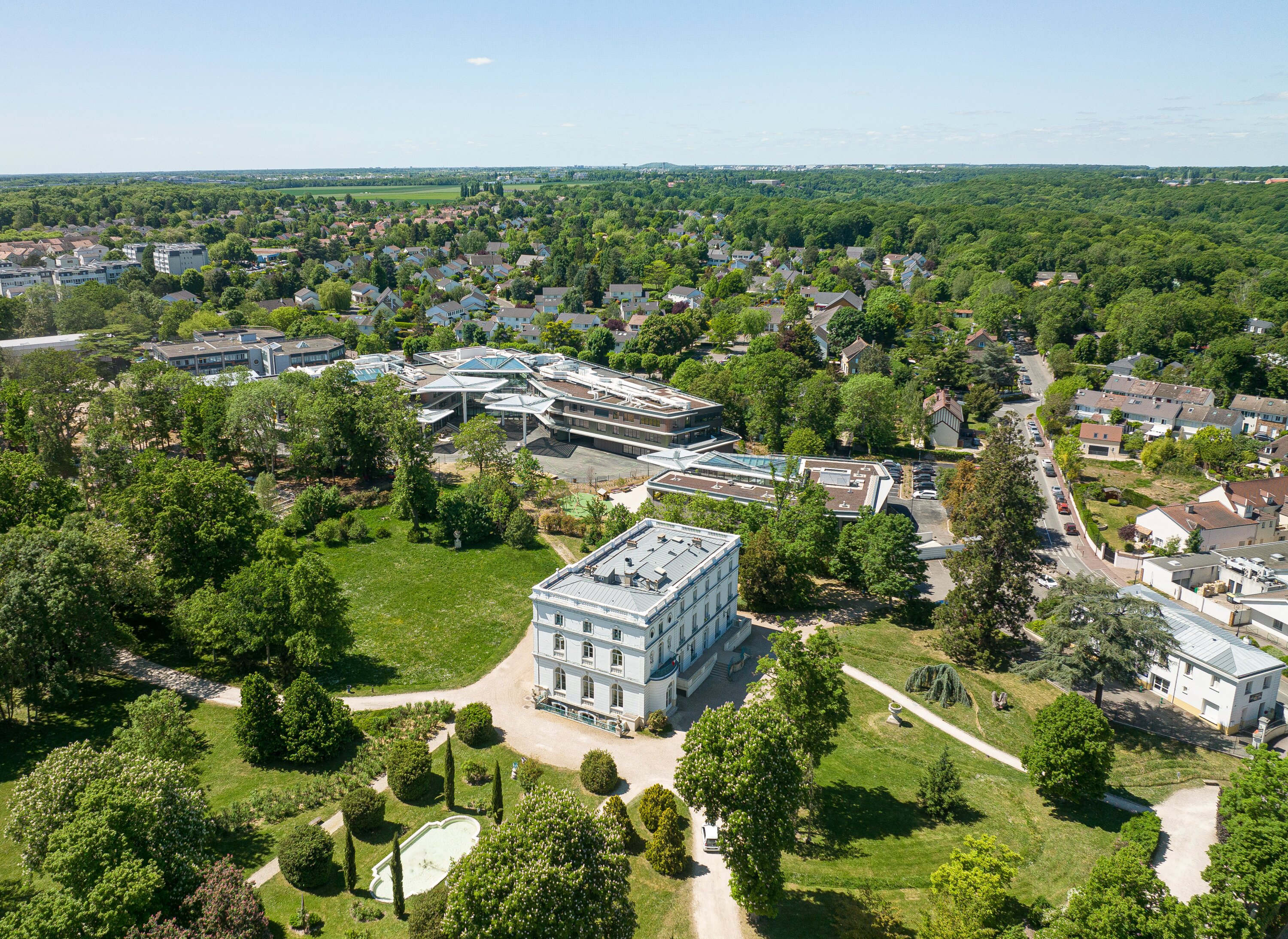 Project by Behnisch Architekturbuero, Lycee Franco Allemand. Aerial view of the campus with surrounding parkland and nearby residential area under a clear sky.
