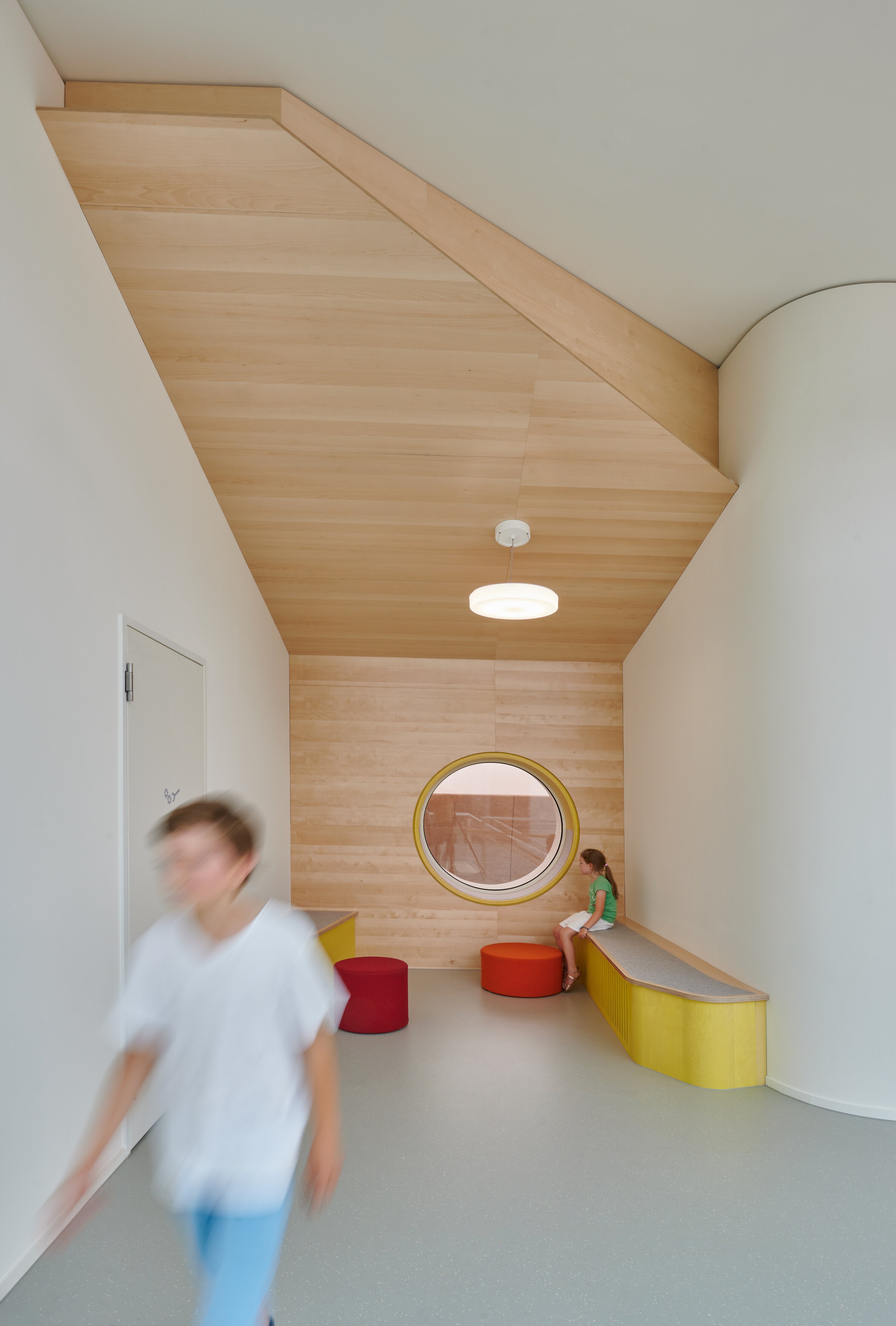 Project by Behnisch Architekturbuero, Gotthard-M&uuml;ller-School Bernhausen. A narrow school hallway with a wooden ceiling, a round window, and a built-in bench; one child is sitting, another is walking past. The seated child is sitting on a yellow cushion. There are also red stools.