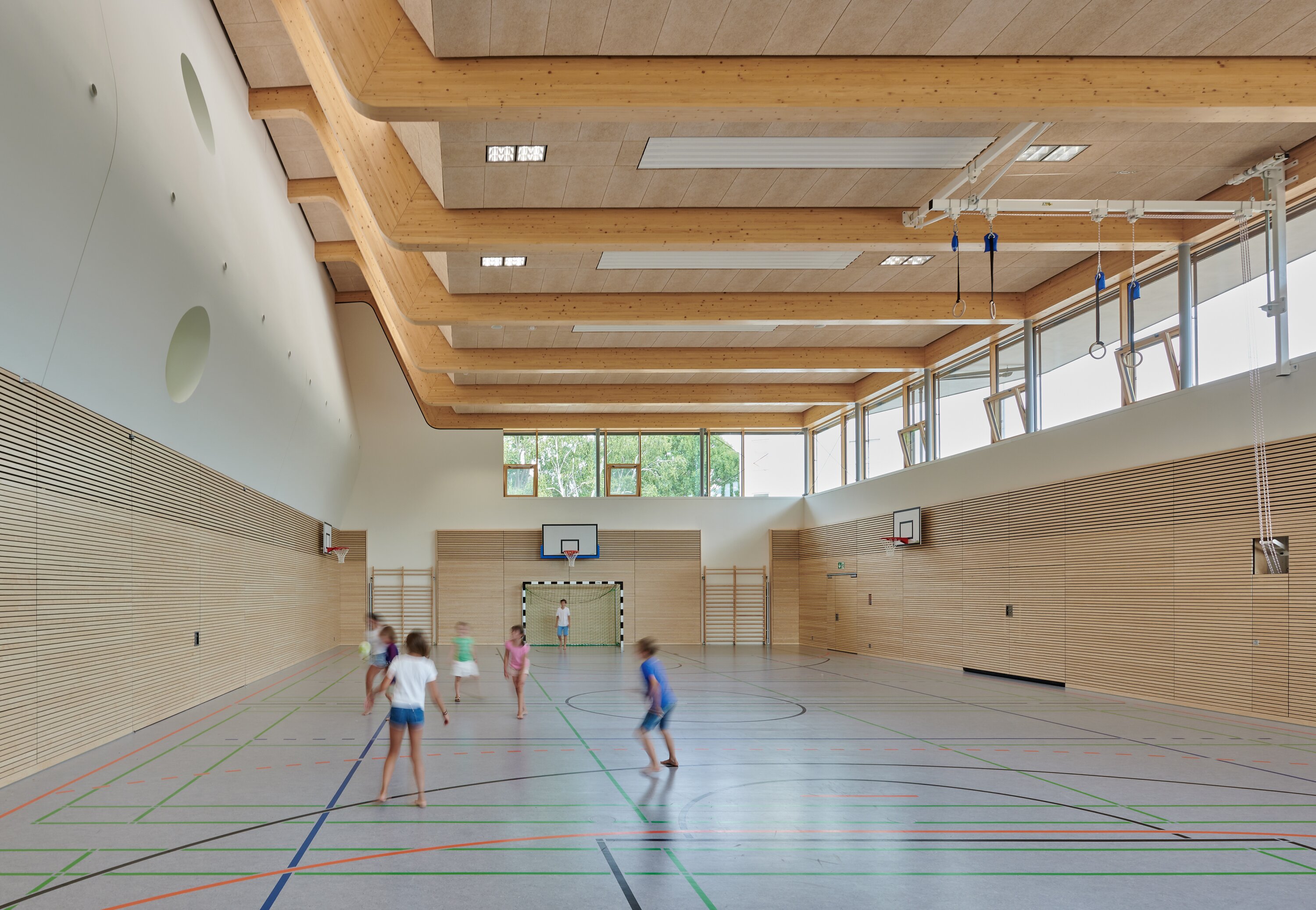 Project by Behnisch Architekturbuero, Gotthard-M&uuml;ller-School Bernhausen. School gym with wooden walls and high ceiling, children running and playing on marked court, basketball hoops and goal at far end.