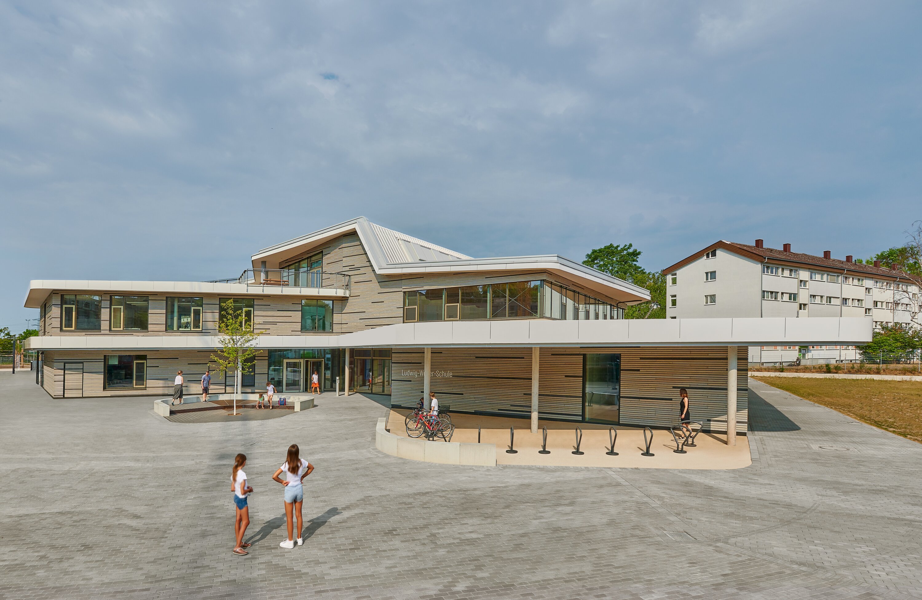 Project by Behnisch Architekturbuero, Gotthard-M&uuml;ller-School Bernhausen. A modern school building with an angular roof and glass facade; students stand and walk in a large paved courtyard. There are several bicycle parking areas in front of the building.