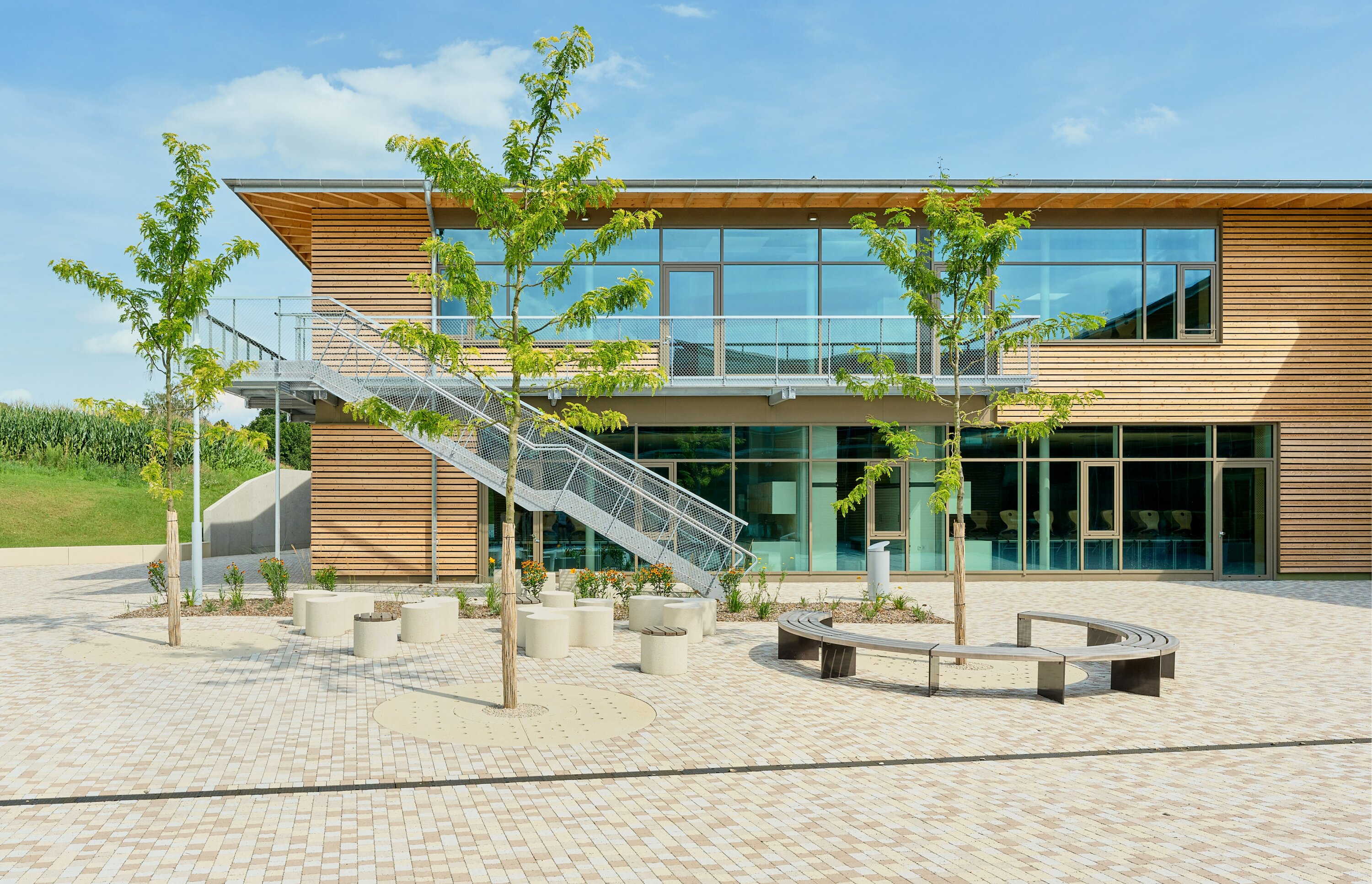 Project by Behnisch Architekturbuero, Public Secondary School Neuburg an der Donau. Two-story school building with wood facade and large windows, exterior stairs, and a paved courtyard with trees, benches, and circular seating.
