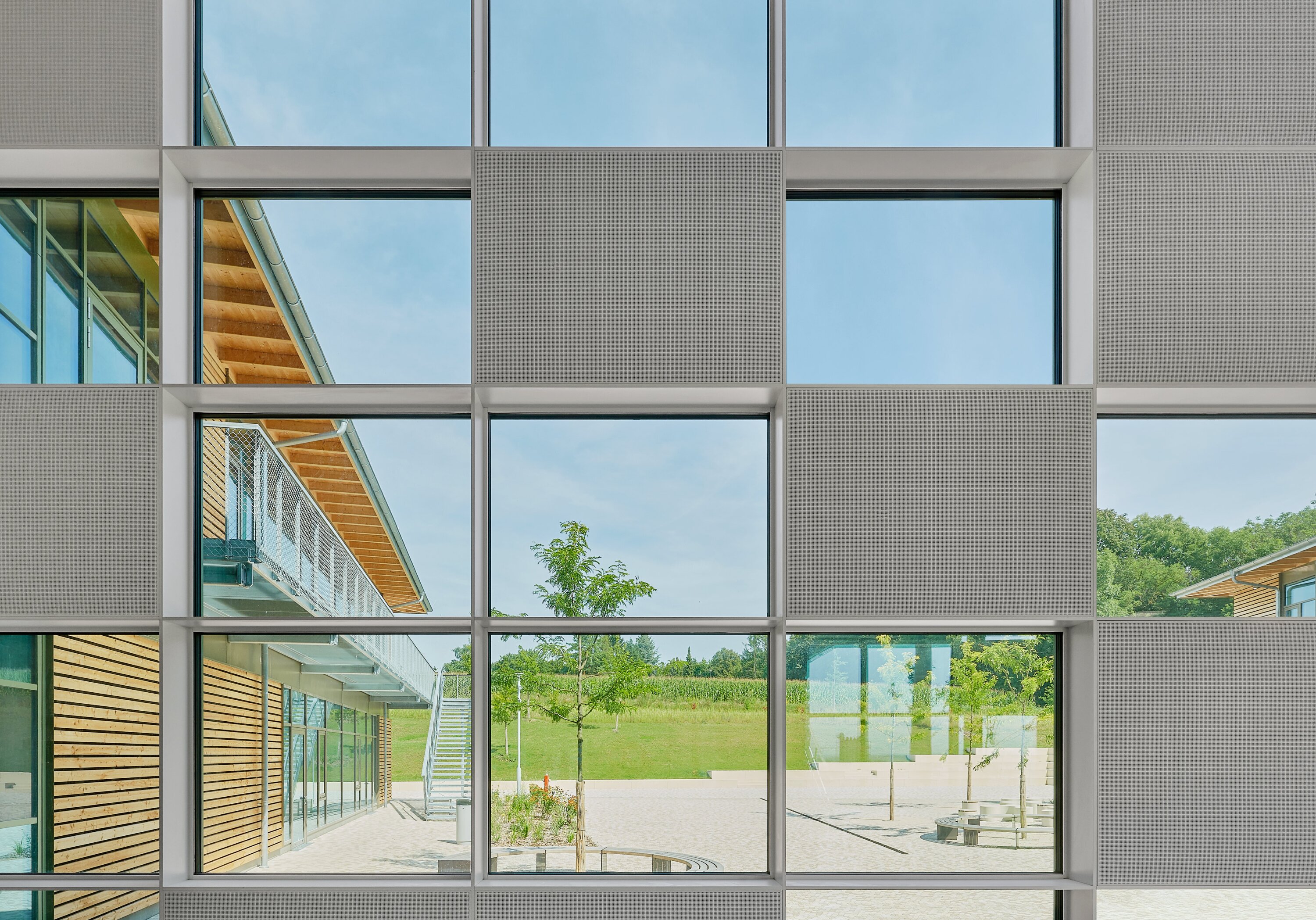 Project by Behnisch Architekturbuero, Public Secondary School Neuburg an der Donau. Grid of large windows with opaque panels, showing view of the school courtyard with trees, benches, and a wood-clad building with exterior stairs.