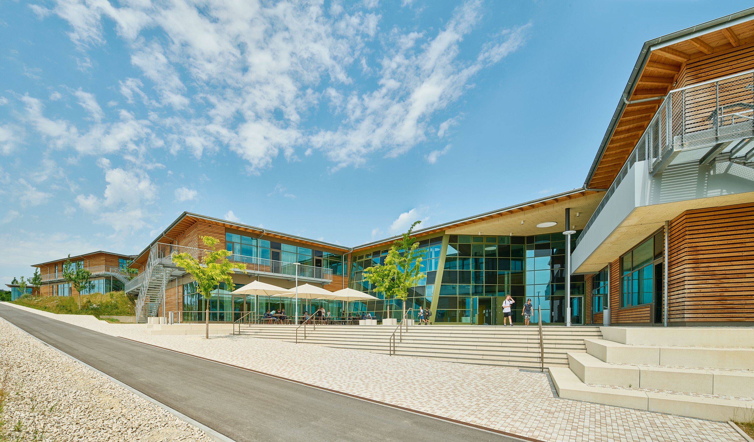 Project by Behnisch Architekturbuero, Public Secondary School Neuburg an der Donau. A modern, single-story building with wood cladding and a large glass facade, featuring outdoor seating with parasols. The building is accessed via spacious external staircases. Several students are standing in front of the entrance.