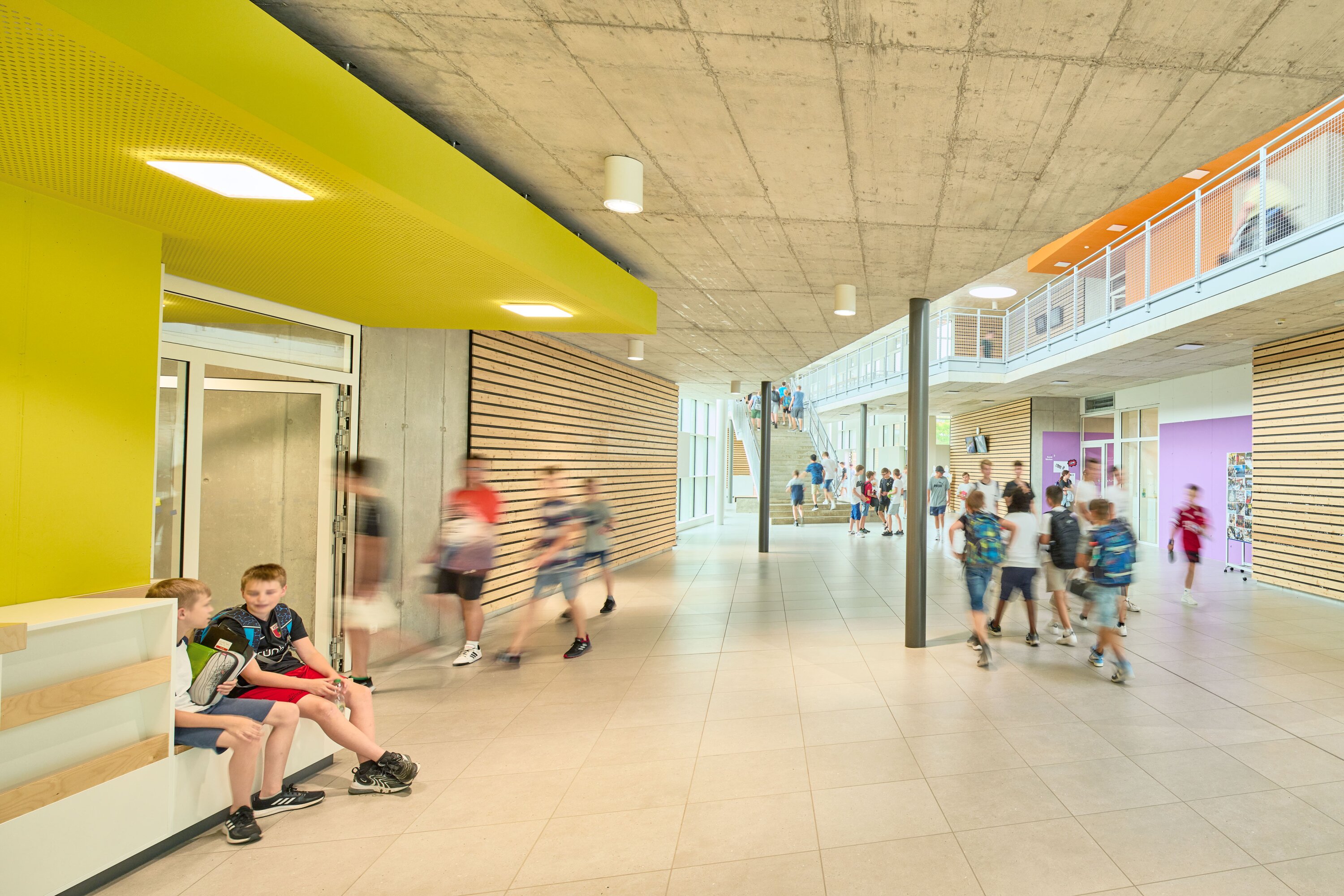 Project by Behnisch Architekturbuero, Public Secondary School Neuburg an der Donau. School corridor with students walking and sitting, yellow ceiling feature, wood wall panels, and stairs leading to an upper-level walkway.