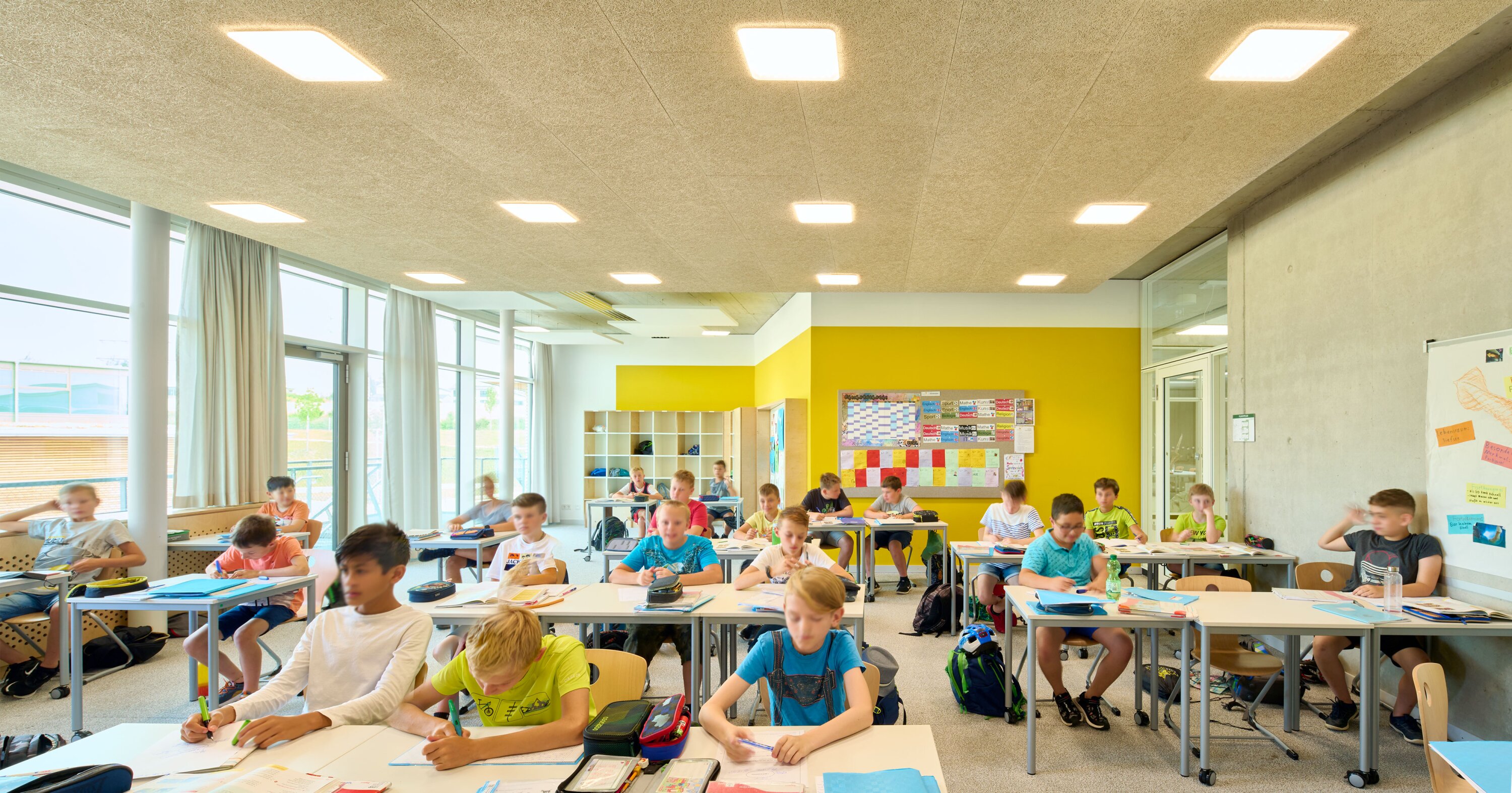 Project by Behnisch Architekturbuero, Public Secondary School Neuburg an der Donau. Classroom with students seated at desks writing, large windows on the left, yellow accent wall, and shelves and board at the back.
